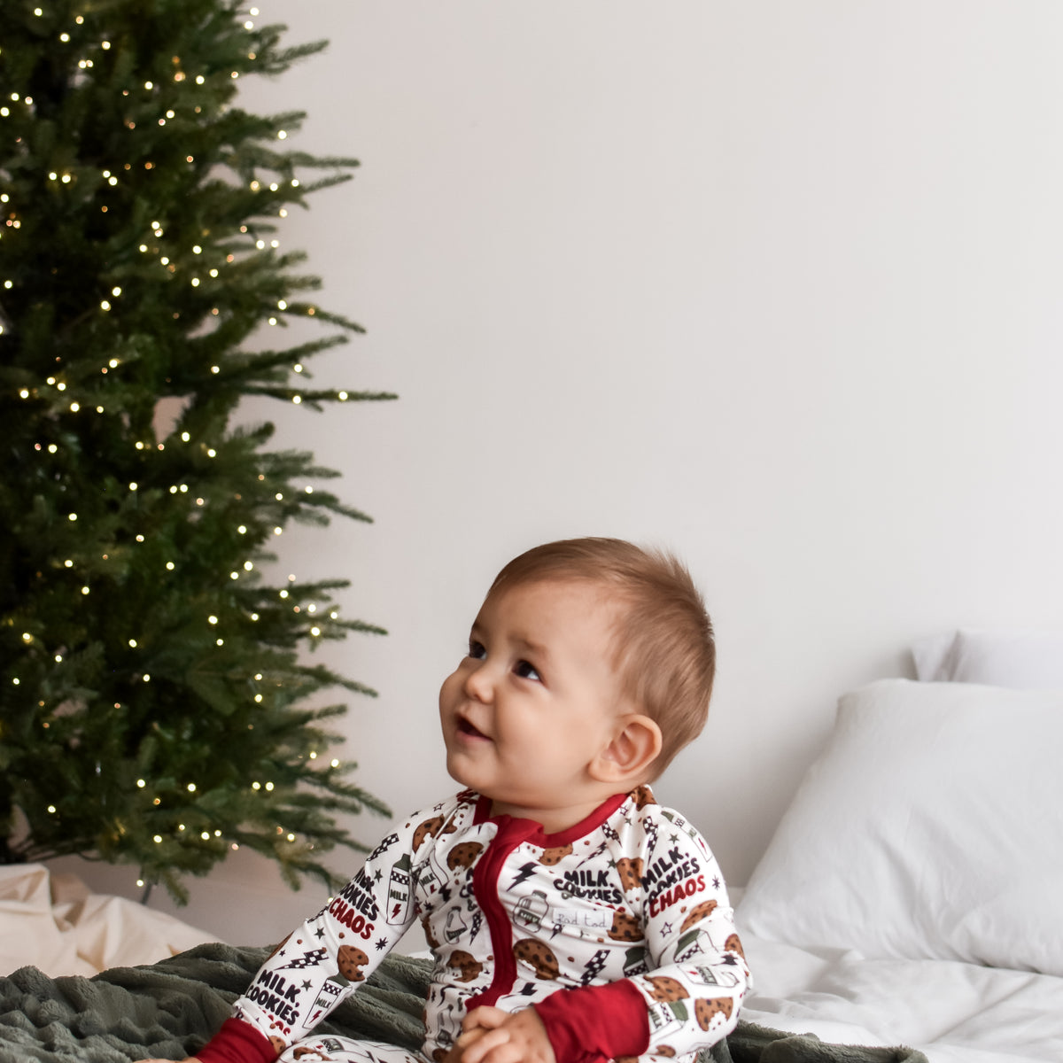 A baby sitting on a textured green blanket in front of a decorated Christmas tree, wearing a white and red holiday print one-piece pajama.