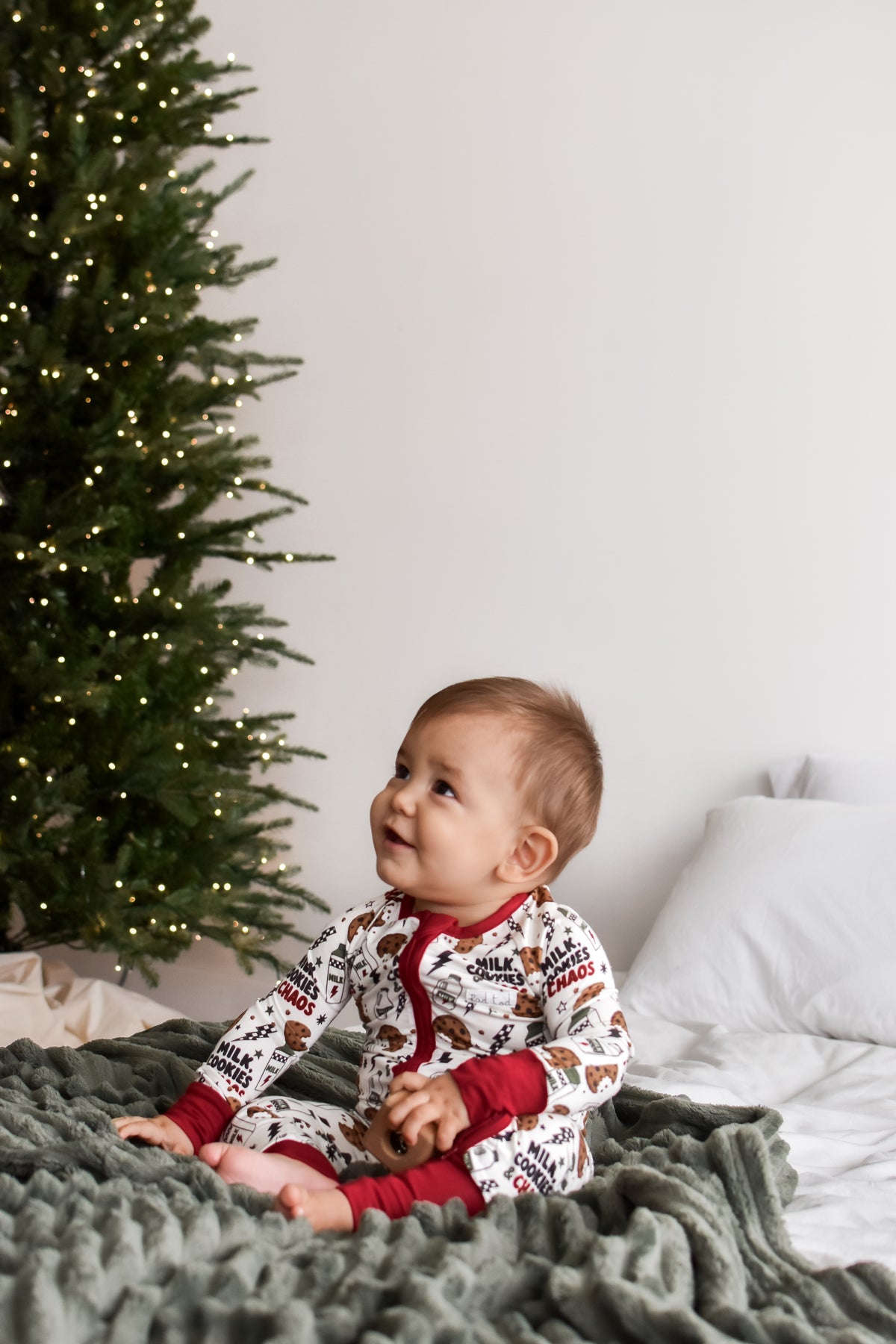 A baby sitting on a textured green blanket in front of a decorated Christmas tree, wearing a white and red holiday print one-piece pajama.