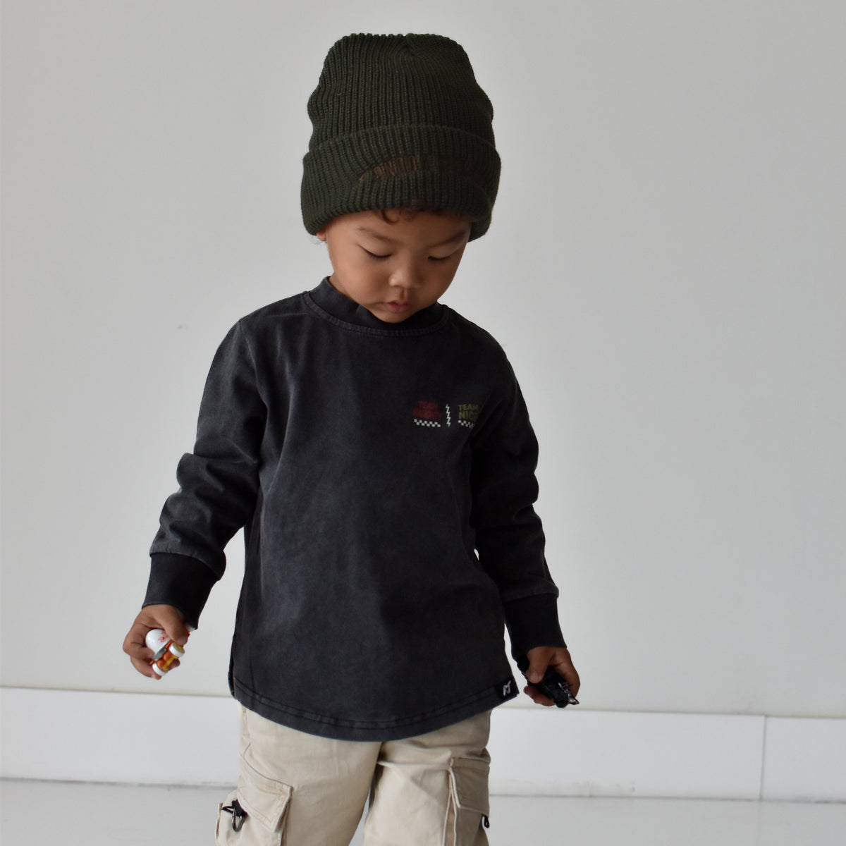 A toddler wearing a dark charcoal long-sleeve shirt with a small chest graphic, beige cargo pants, and a dark olive green knit beanie, standing against a minimalist light gray background.