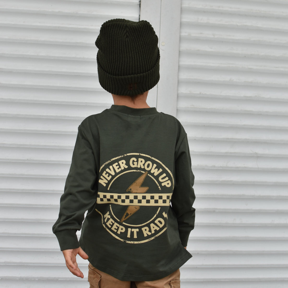 A child viewed from behind wearing an olive green long-sleeve shirt with a tan graphic print on the back, paired with a black knit beanie and tan cargo pants, standing in front of a white corrugated metal wall.