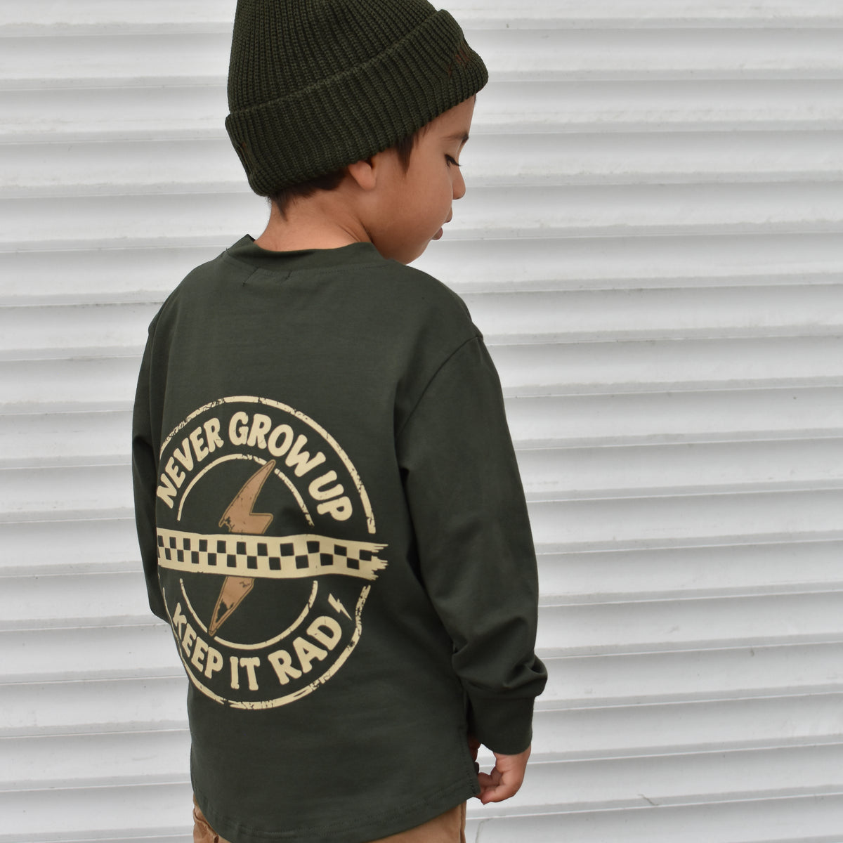 A child wearing an olive green long-sleeve shirt with a large graphic print on the back, paired with a dark green beanie and tan cargo pants, standing in front of a white corrugated metal wall.
