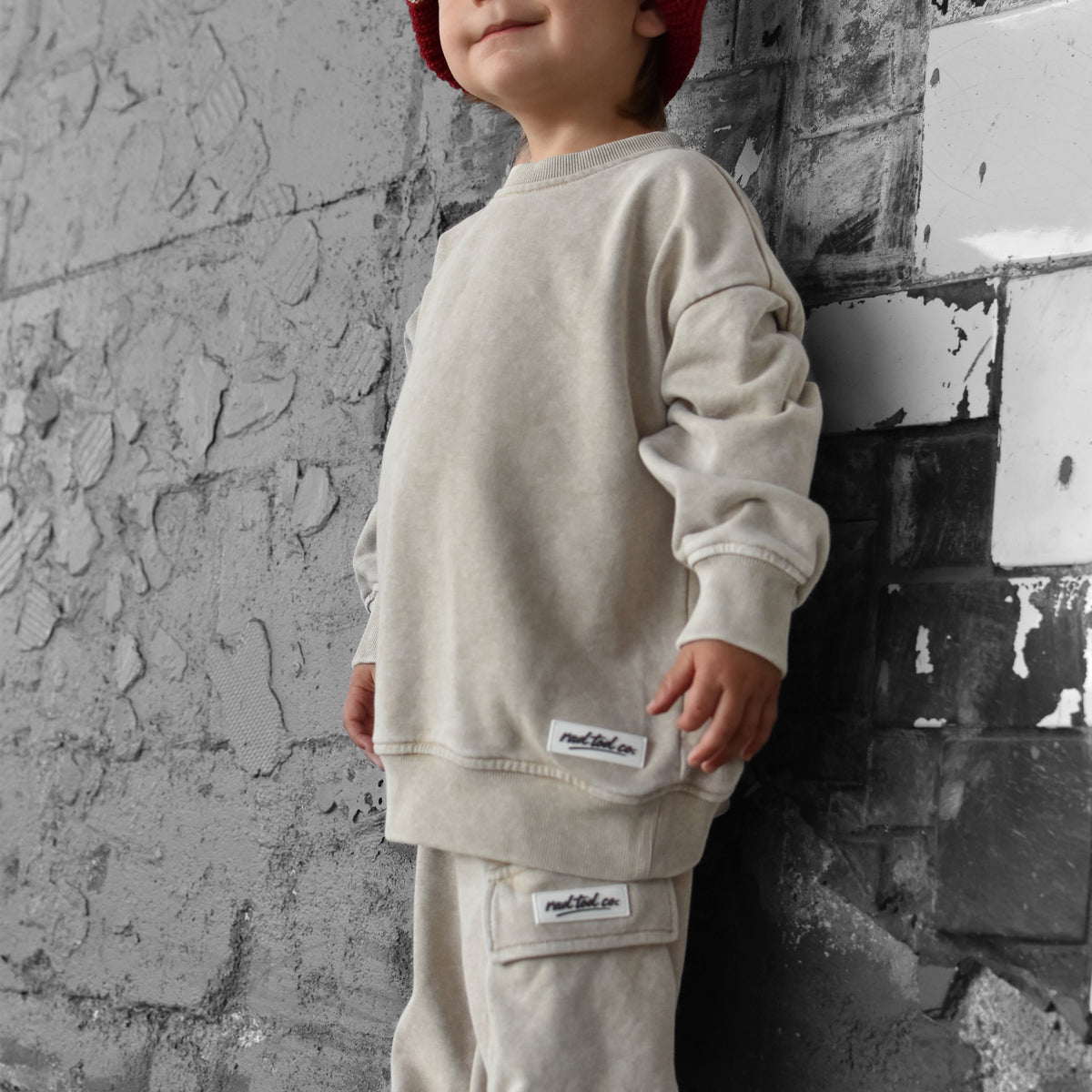A toddler stands against a textured gray brick wall wearing a cream-colored sweatshirt and matching cargo pants, paired with a red knit beanie.