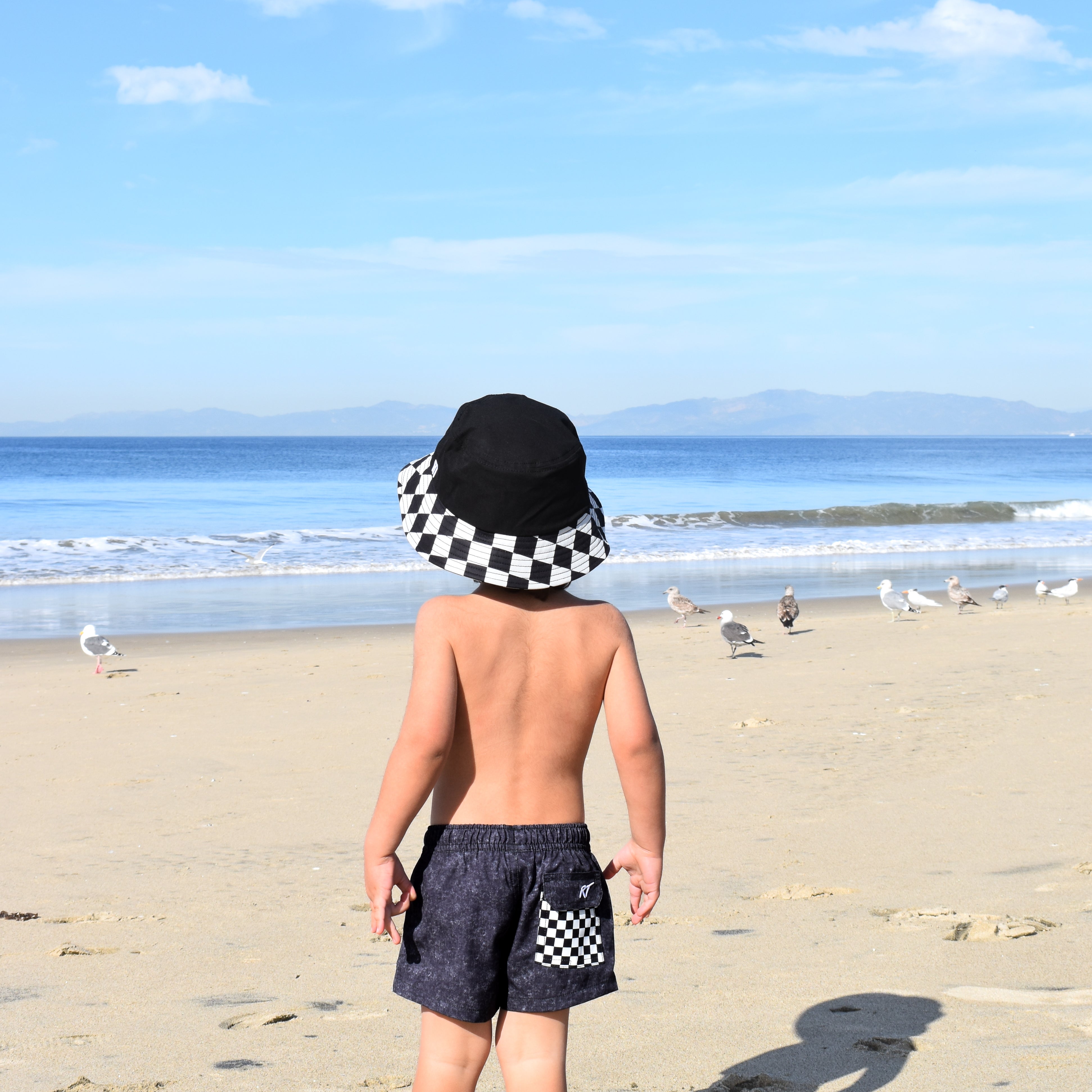 Child standing on a beach wearing a checkered hat and shorts, looking at the ocean.