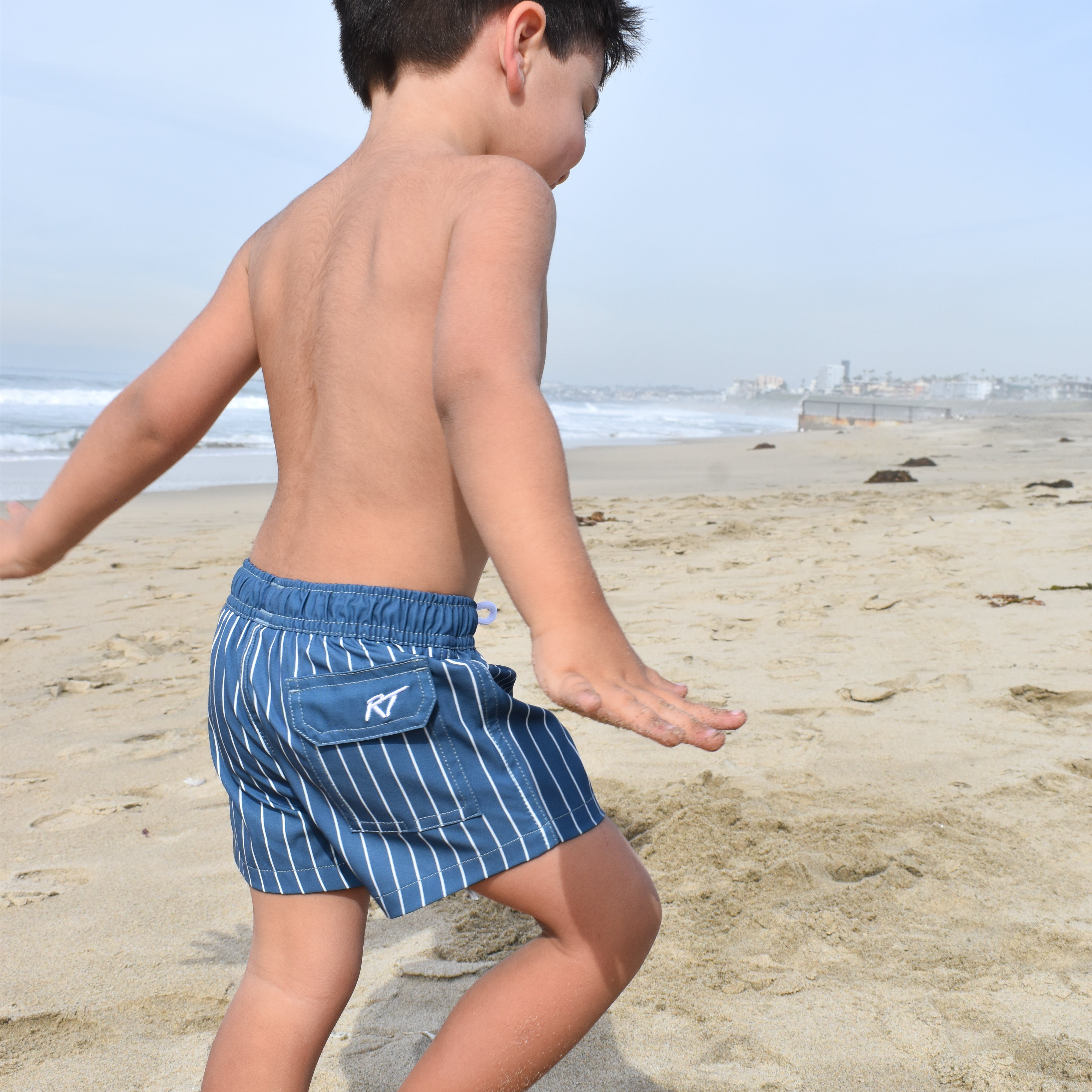 Child wearing blue striped swim trunks on a sandy beach with ocean in the background
