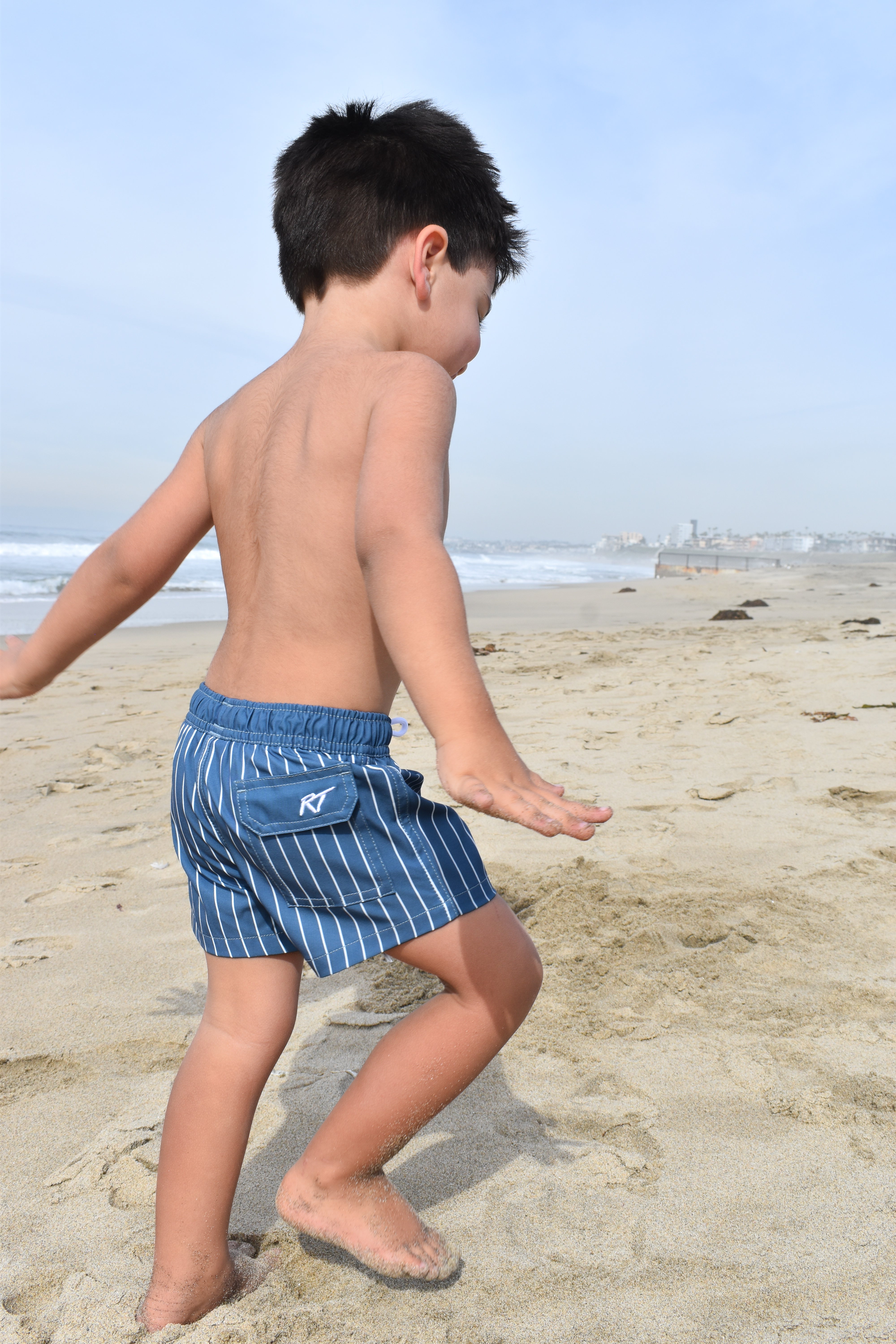 Child wearing blue striped swim trunks on a sandy beach with ocean in the background