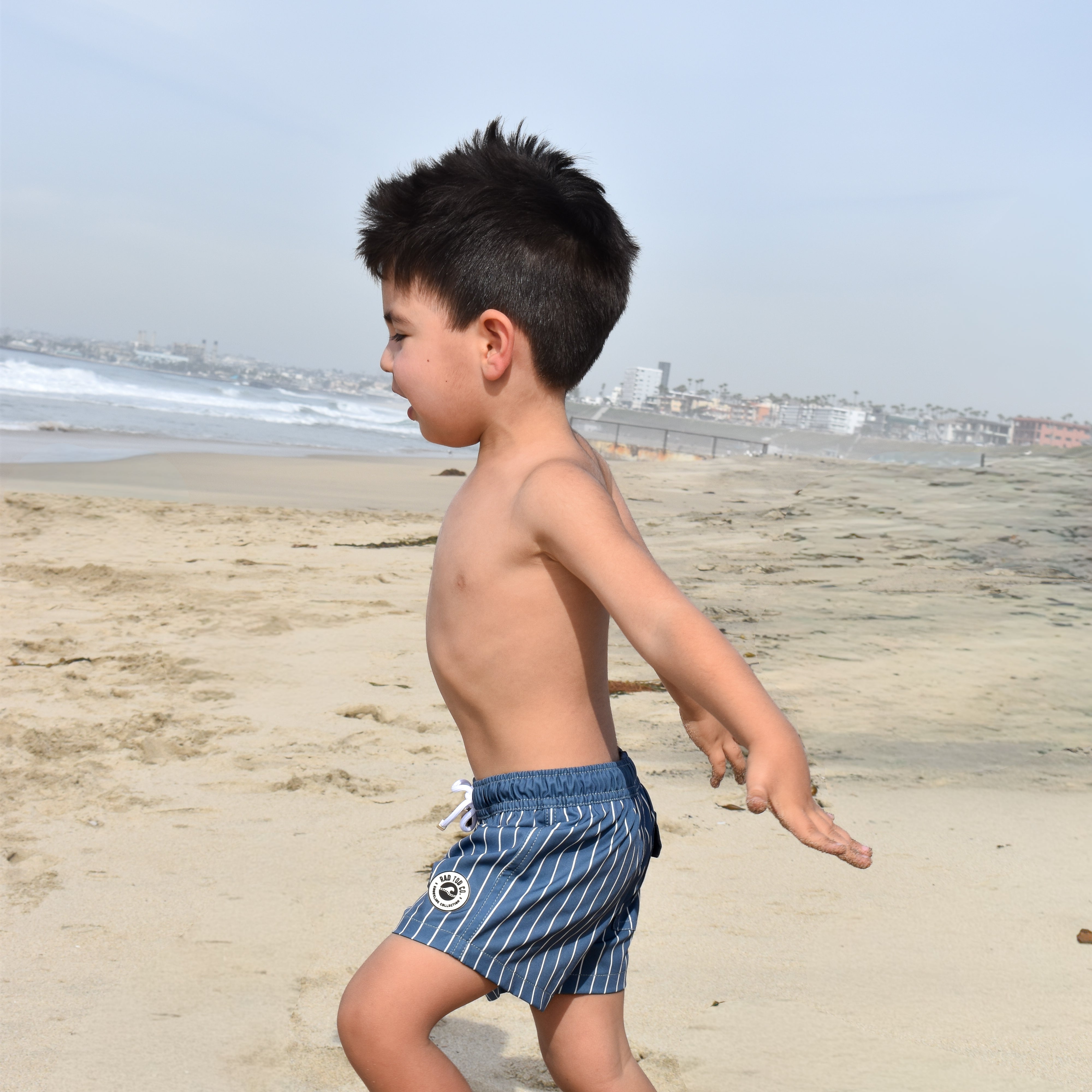 Child in striped rad toddler swim shorts walking on a sandy beach with ocean and sky in the background