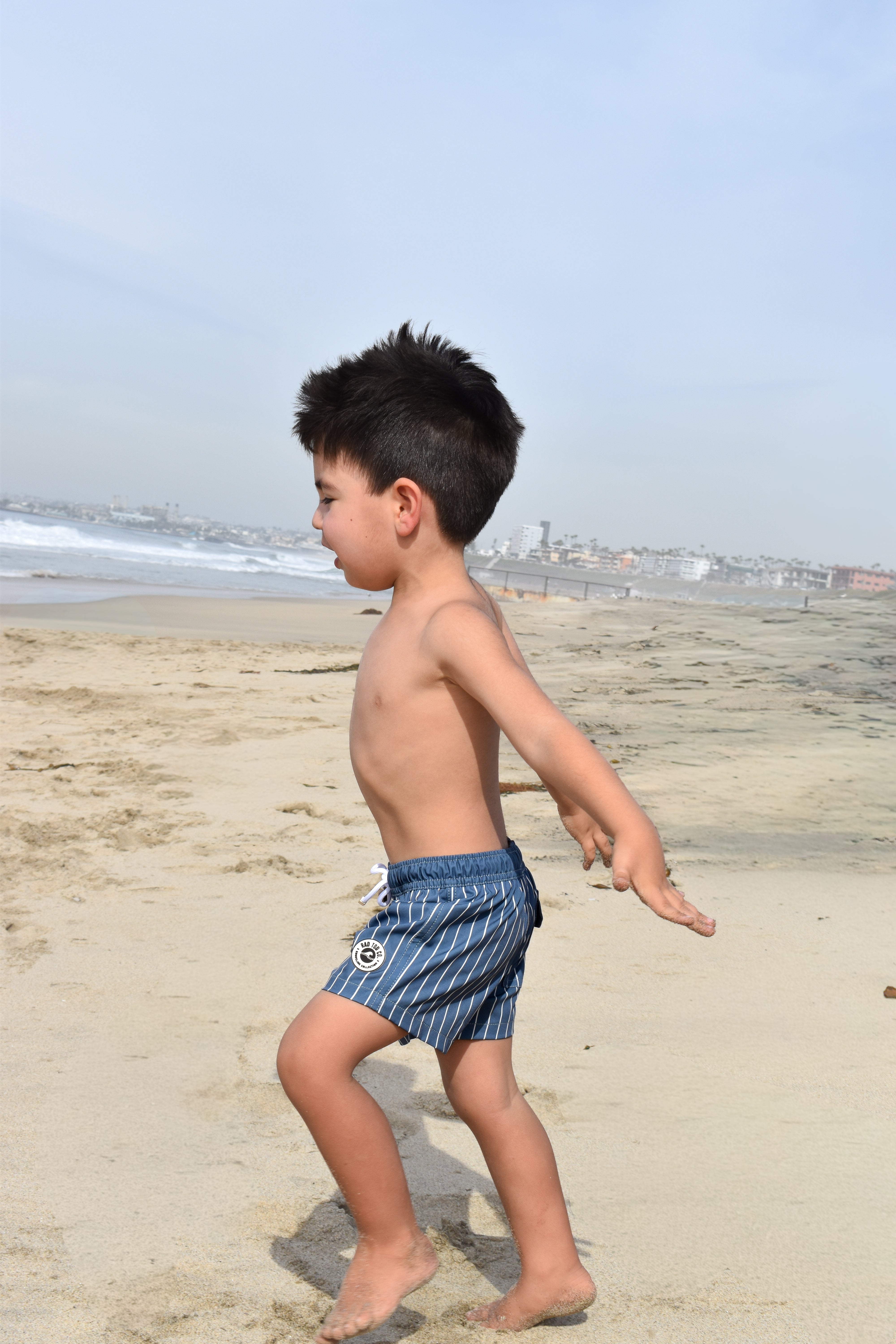 Child in striped rad toddler swim shorts walking on a sandy beach with ocean and sky in the background