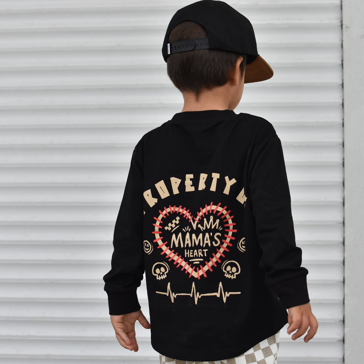 A child wearing a black long-sleeve shirt with a large red and gold heart graphic and the words 'PROPERTY OF MAMA'S HEART' on the back, paired with beige-and-white checkered pants and a black cap, standing in front of a white corrugated metal wall.