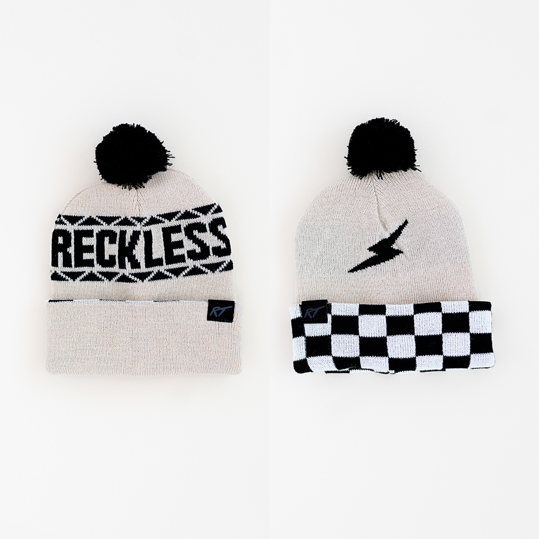 Two reversible toddler beanies, one with a 'RECKLESS' text pattern and the other with a lightning bolt design, both featuring a black pom-pom and a black and white checkered pattern around the base.