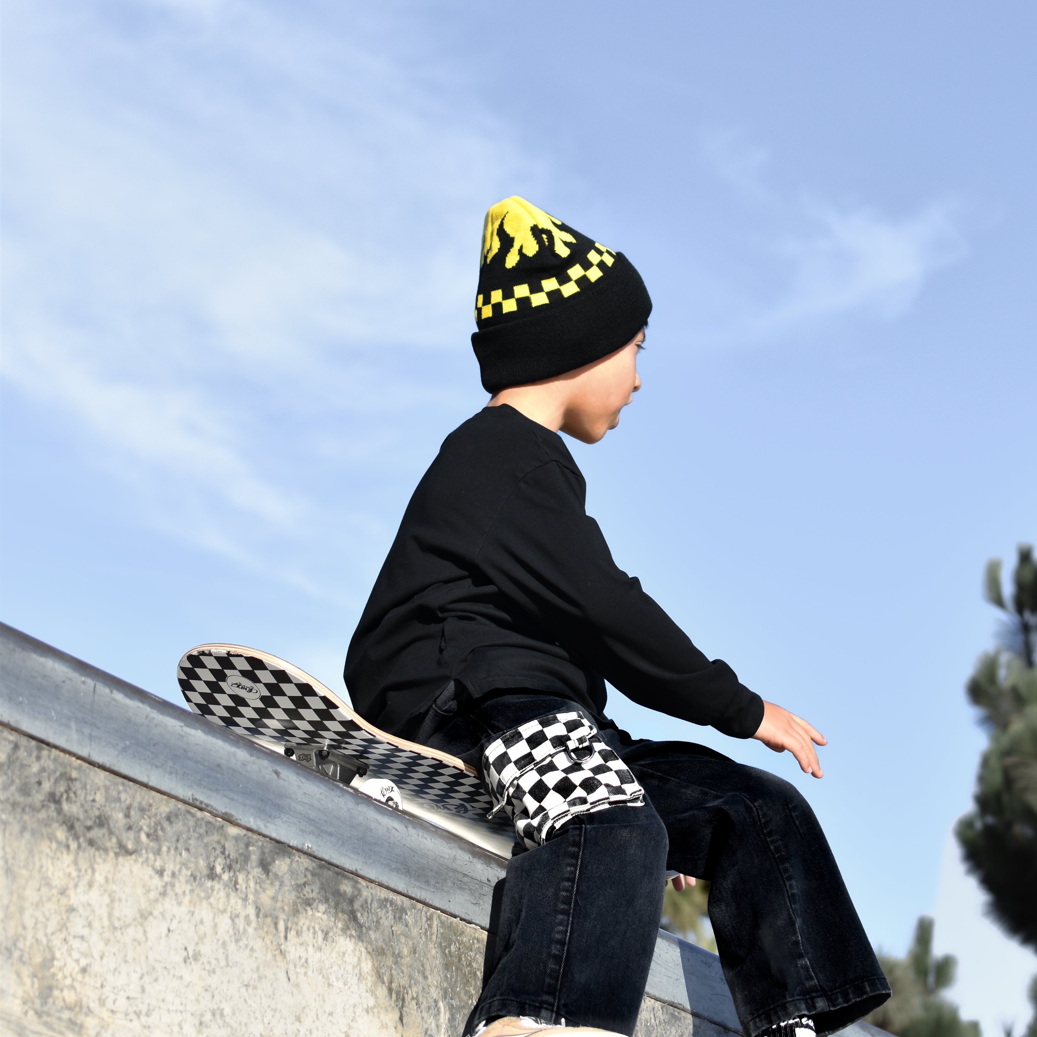 A child wearing a black beanie with yellow details, a black long-sleeve top, and dark jeans sits on the edge of a concrete skate ramp under a blue sky. The child is wearing Vans sneakers and has a Vans skateboard resting on the ramp behind them.
