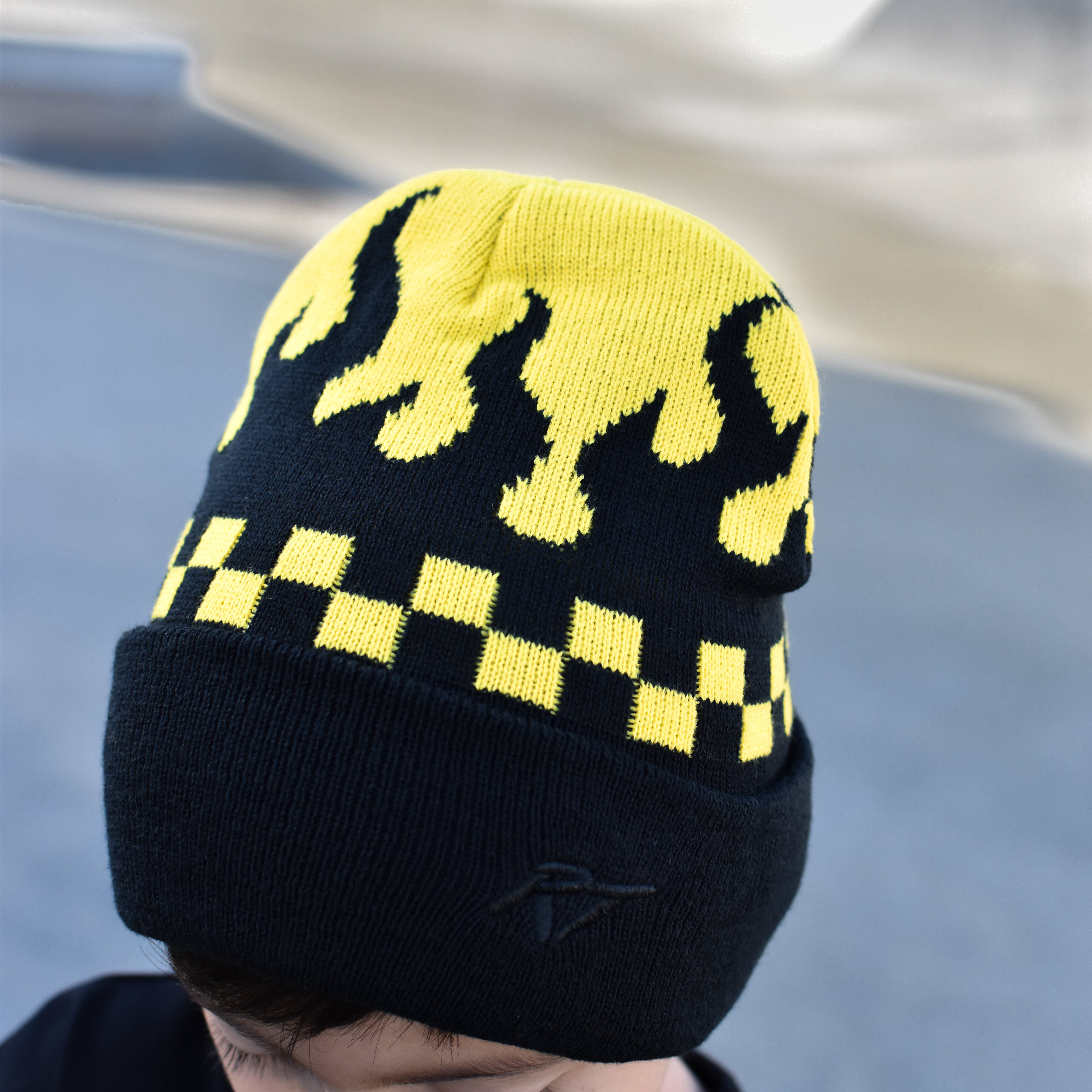 A child wearing a black knit beanie with a yellow flame pattern and a yellow-and-black checkered band, photographed outdoors with a blurred skatepark background.