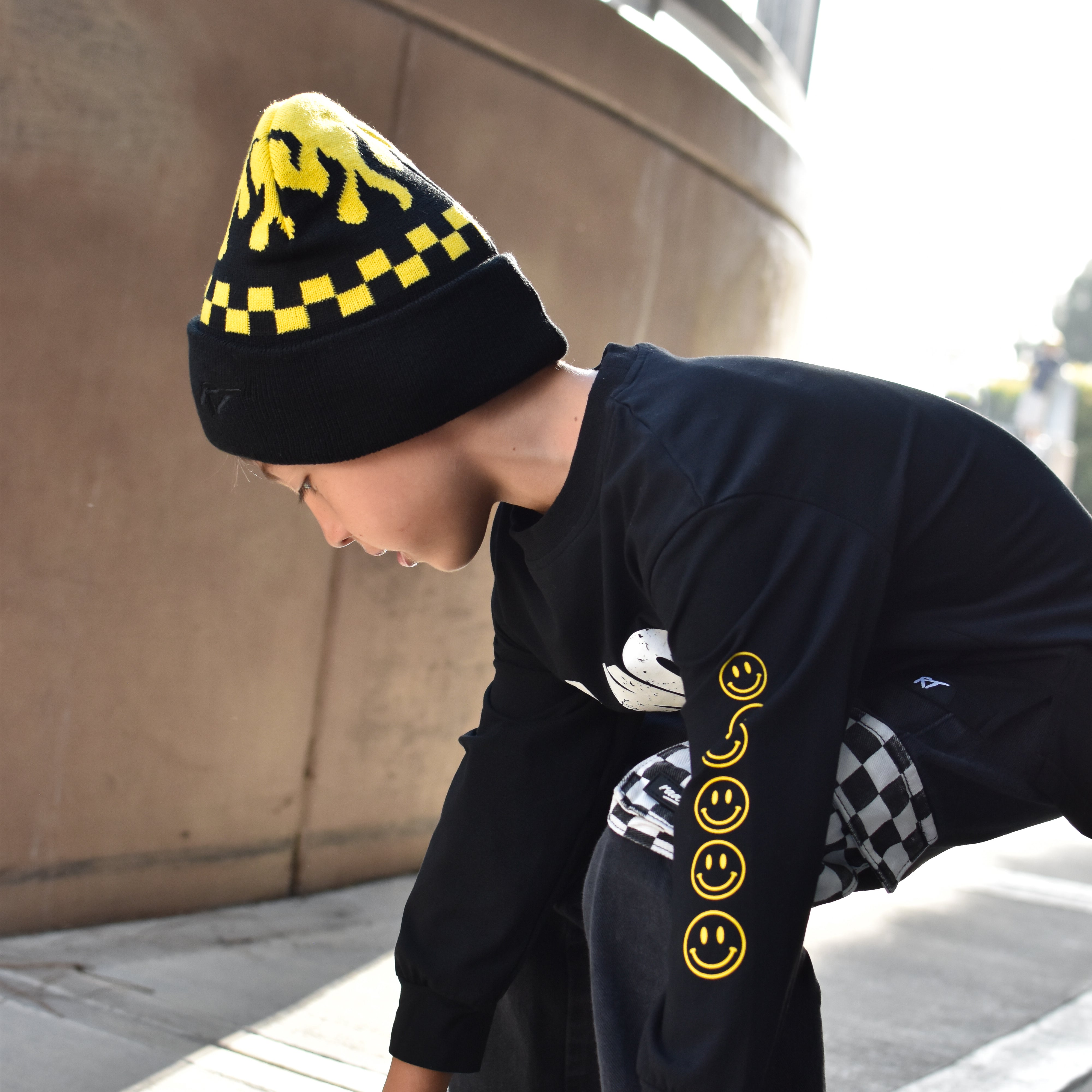 A child wearing a black beanie with yellow graphic details, a black long-sleeve shirt with yellow smiley face graphics on the sleeve, and black pants, crouching on a skateboard outdoors.