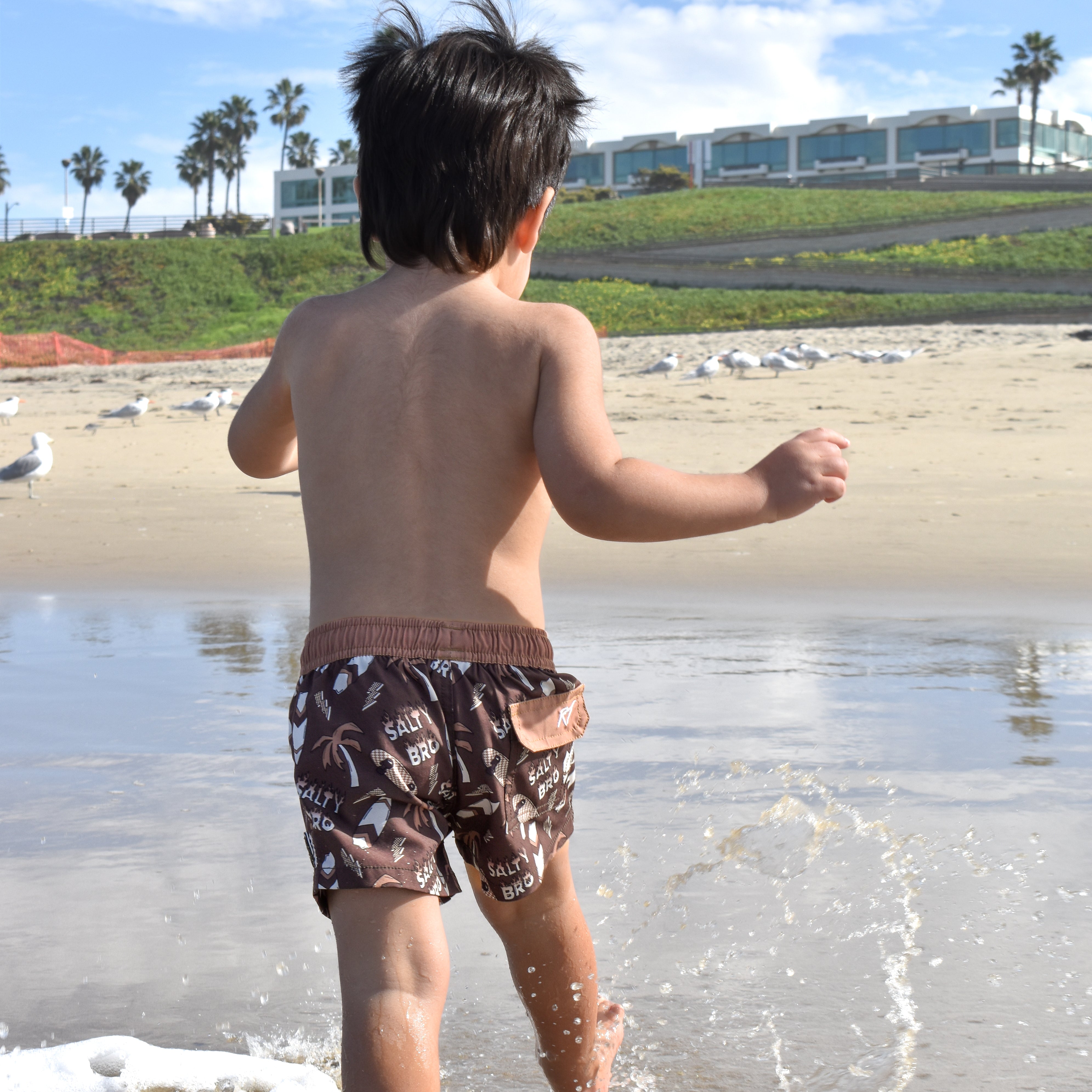 Child playing in shallow water at the beach while wearing Rad Toddler swimmies with palm trees and buildings in the background.