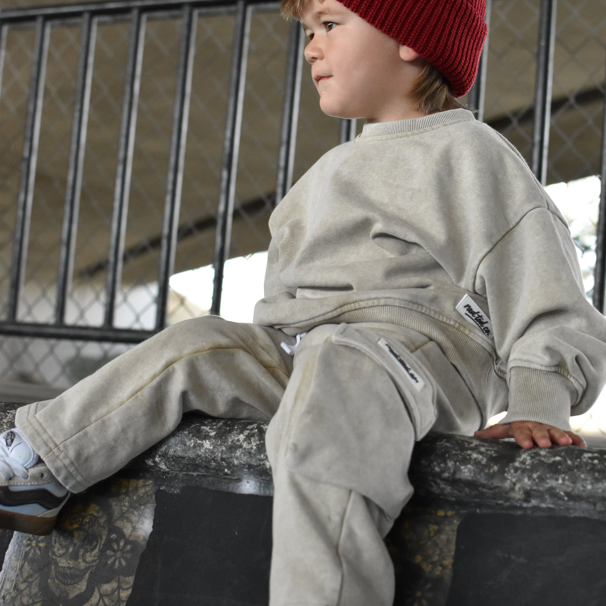 A child wearing a red knit beanie with a small white logo, a light beige sweatshirt, and matching beige pants, sitting on a concrete ledge in an outdoor setting.
