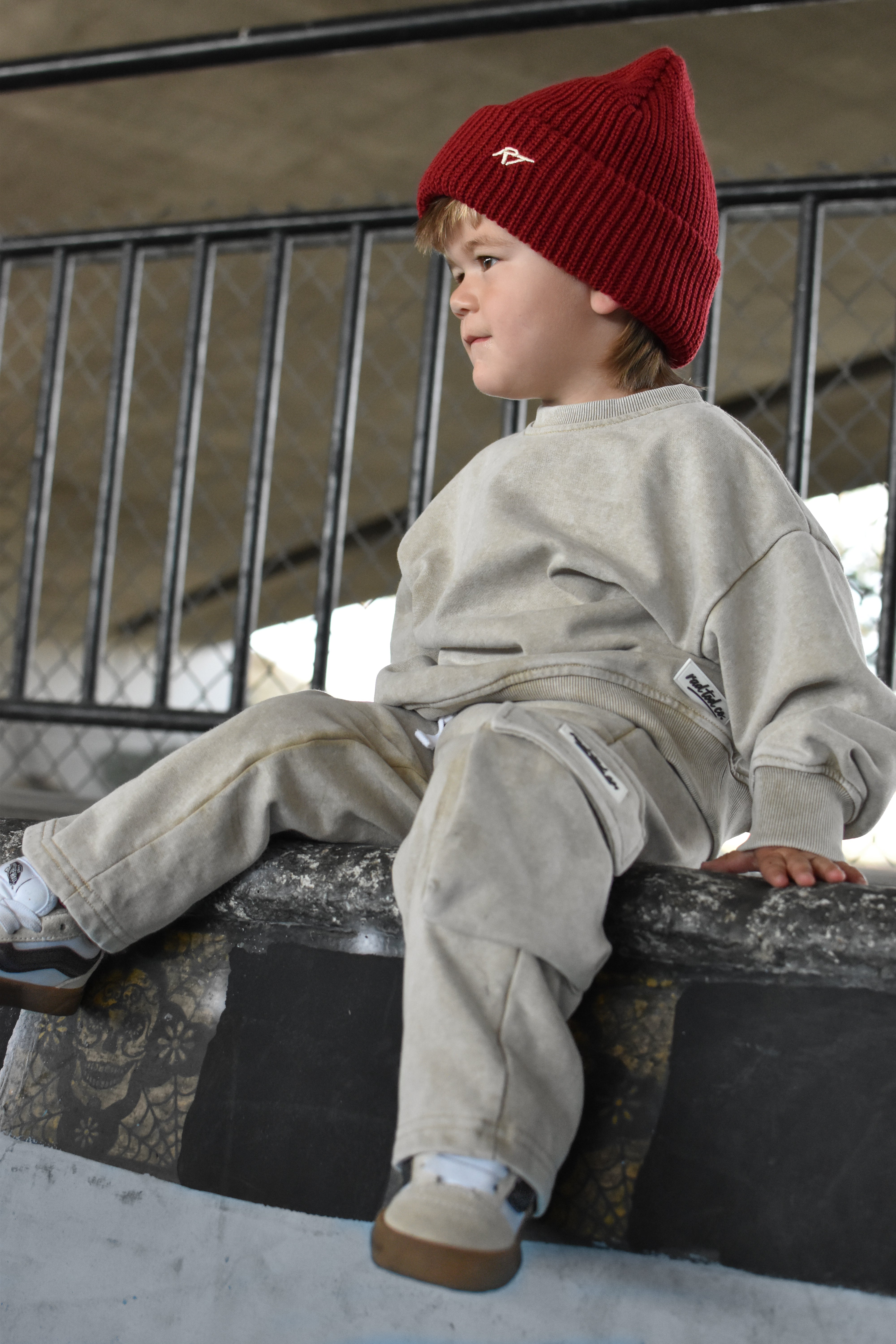 A child wearing a red knit beanie with a small white logo, a light beige sweatshirt, and matching beige pants, sitting on a concrete ledge in an outdoor setting.