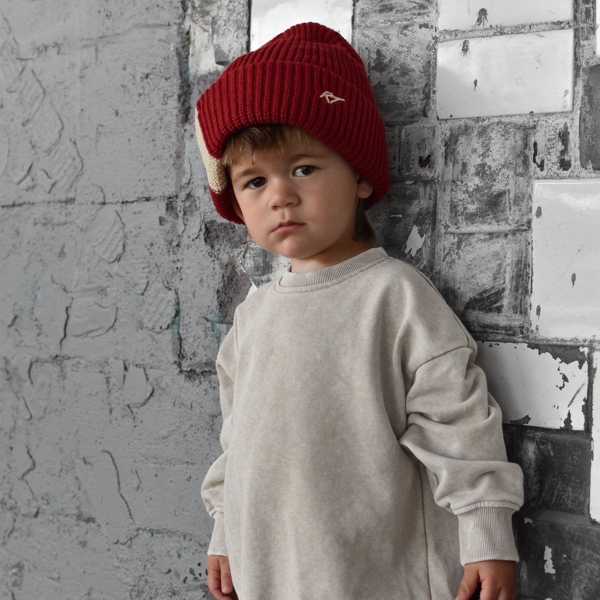 A young child wearing a cream-colored sweatshirt and matching pants with a red knit beanie, standing against a textured gray brick wall.