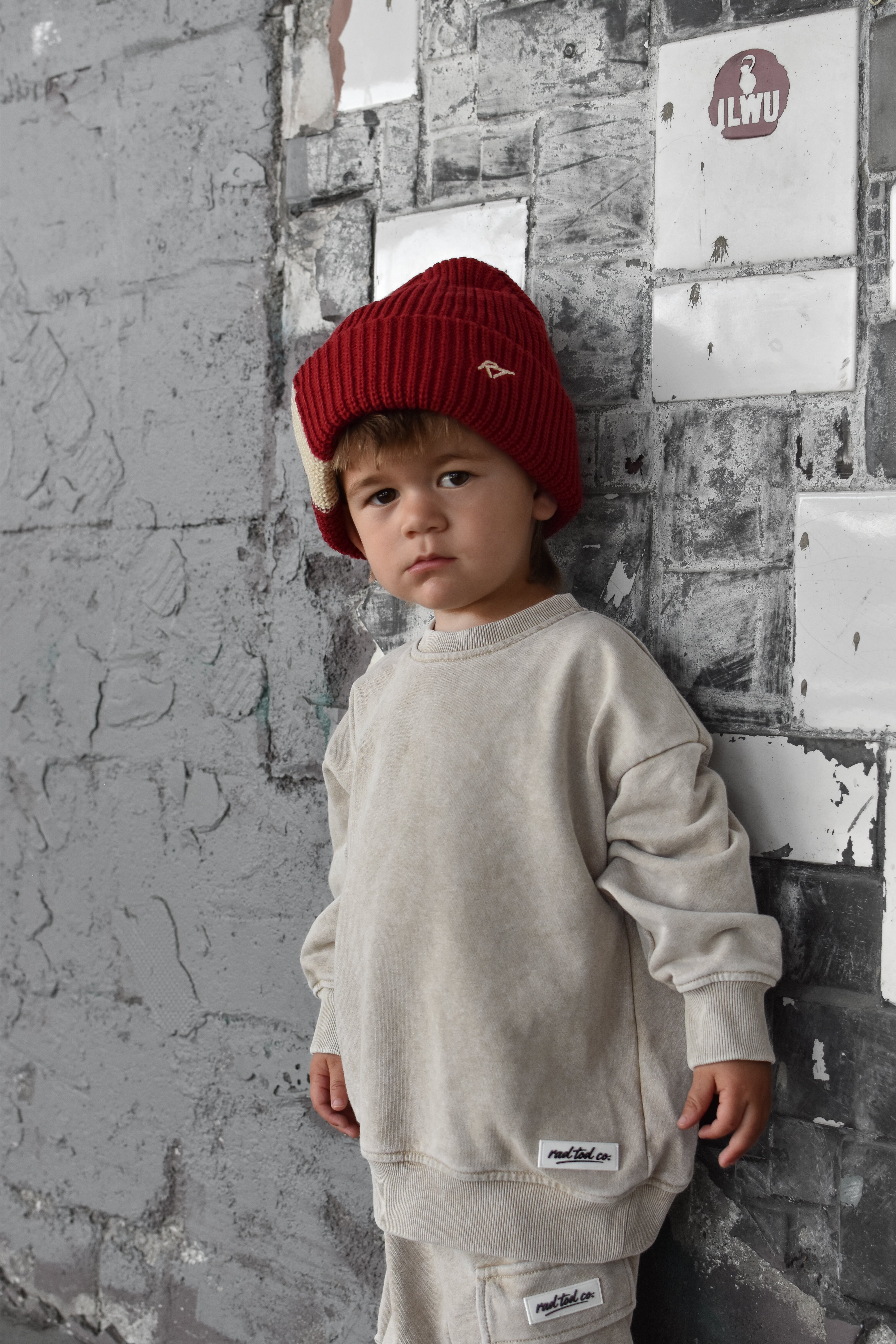 A young child wearing a cream-colored sweatshirt and matching pants with a red knit beanie, standing against a textured gray brick wall.