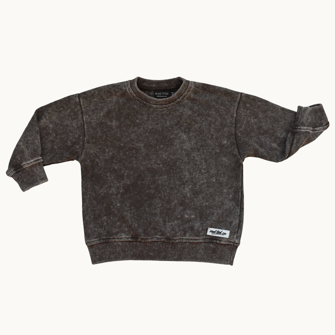 A dark brown acid-wash crewneck sweatshirt with rolled cuffs and a small white brand patch on the lower hem, displayed flat against a light cream background.