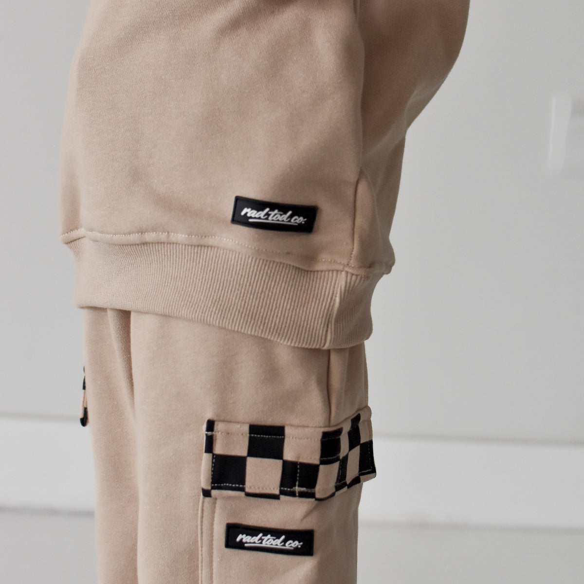 A close-up of a person wearing beige cargo sweatpants with black-and-white checkerboard accents on the pocket trim and cuffs, plus matching beige sweatshirt shown at the top.