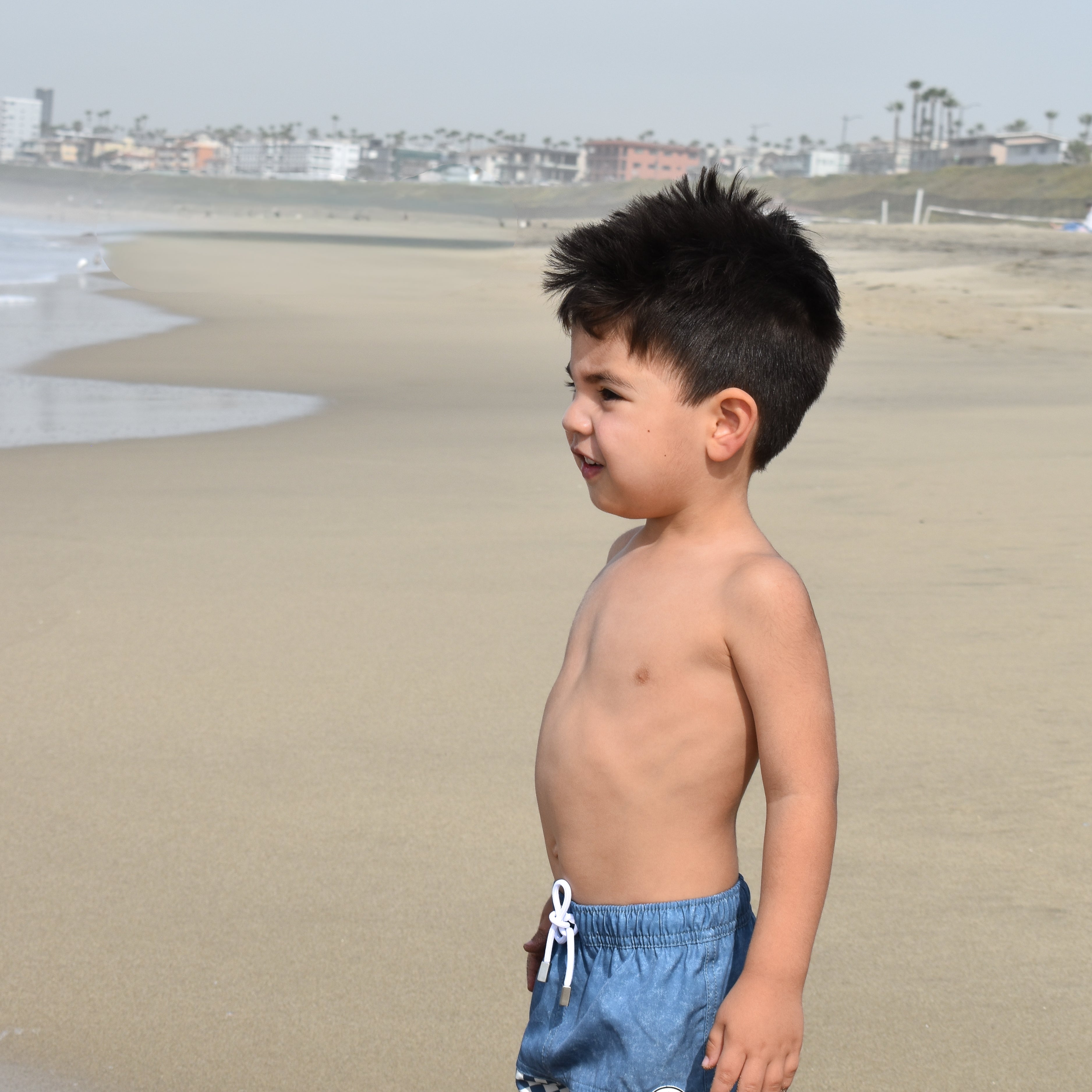 Young boy in blue checkered swim shorts by rad toddler standing on a beach.
