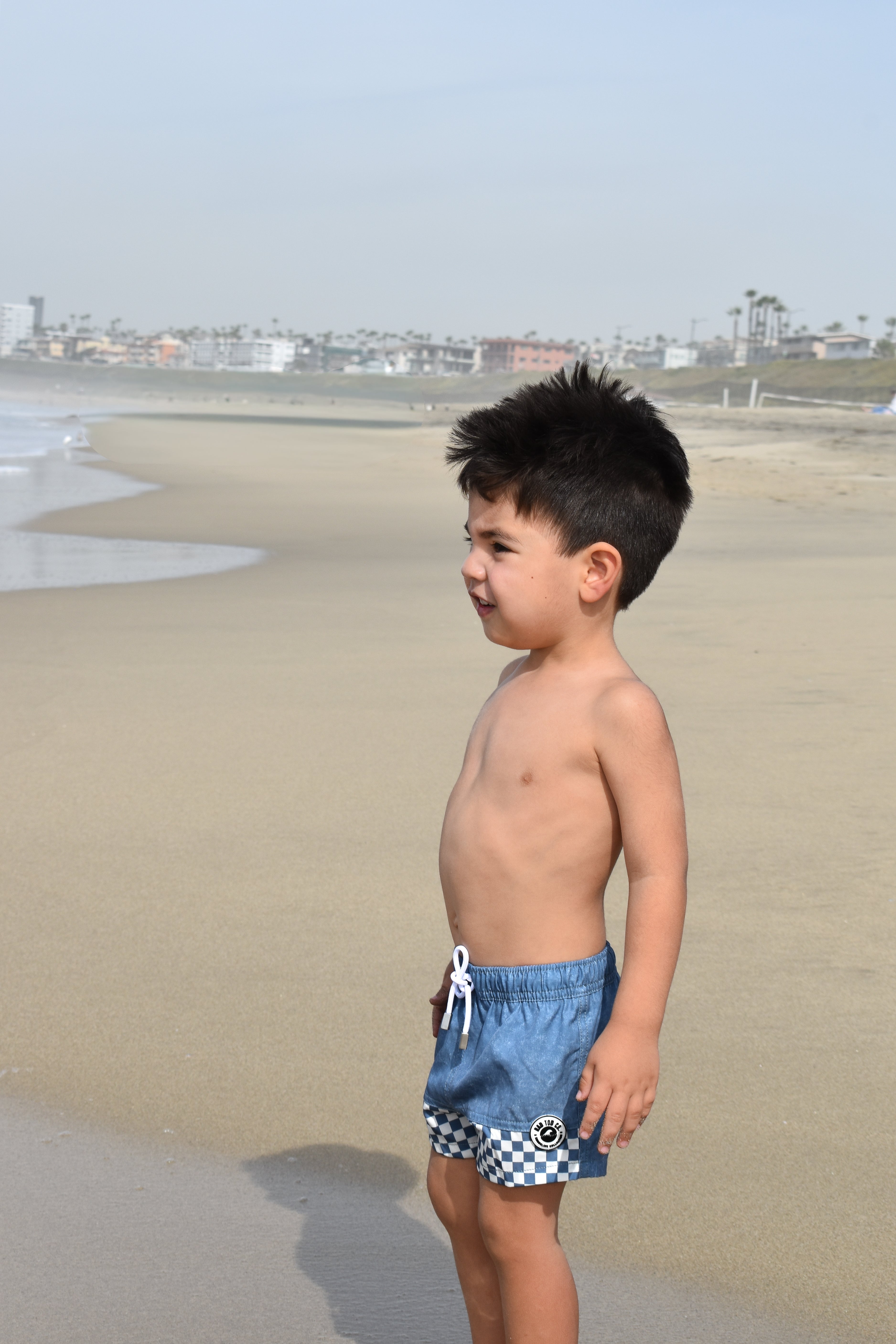 Young boy in blue checkered swim shorts by rad toddler standing on a beach.