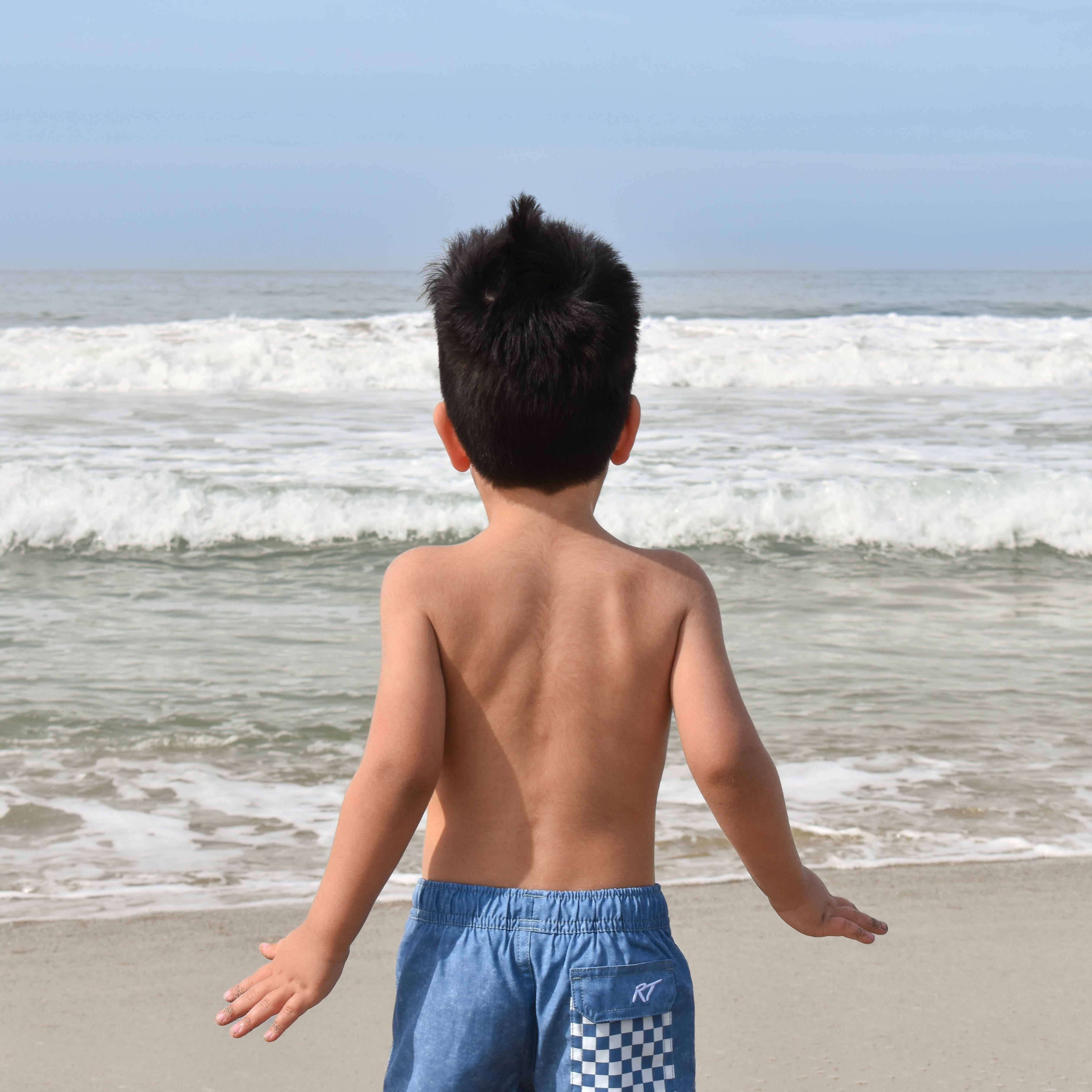 Child wearing blue checkered acid wash shorts standing on a beach with ocean waves in the background by rad toddler