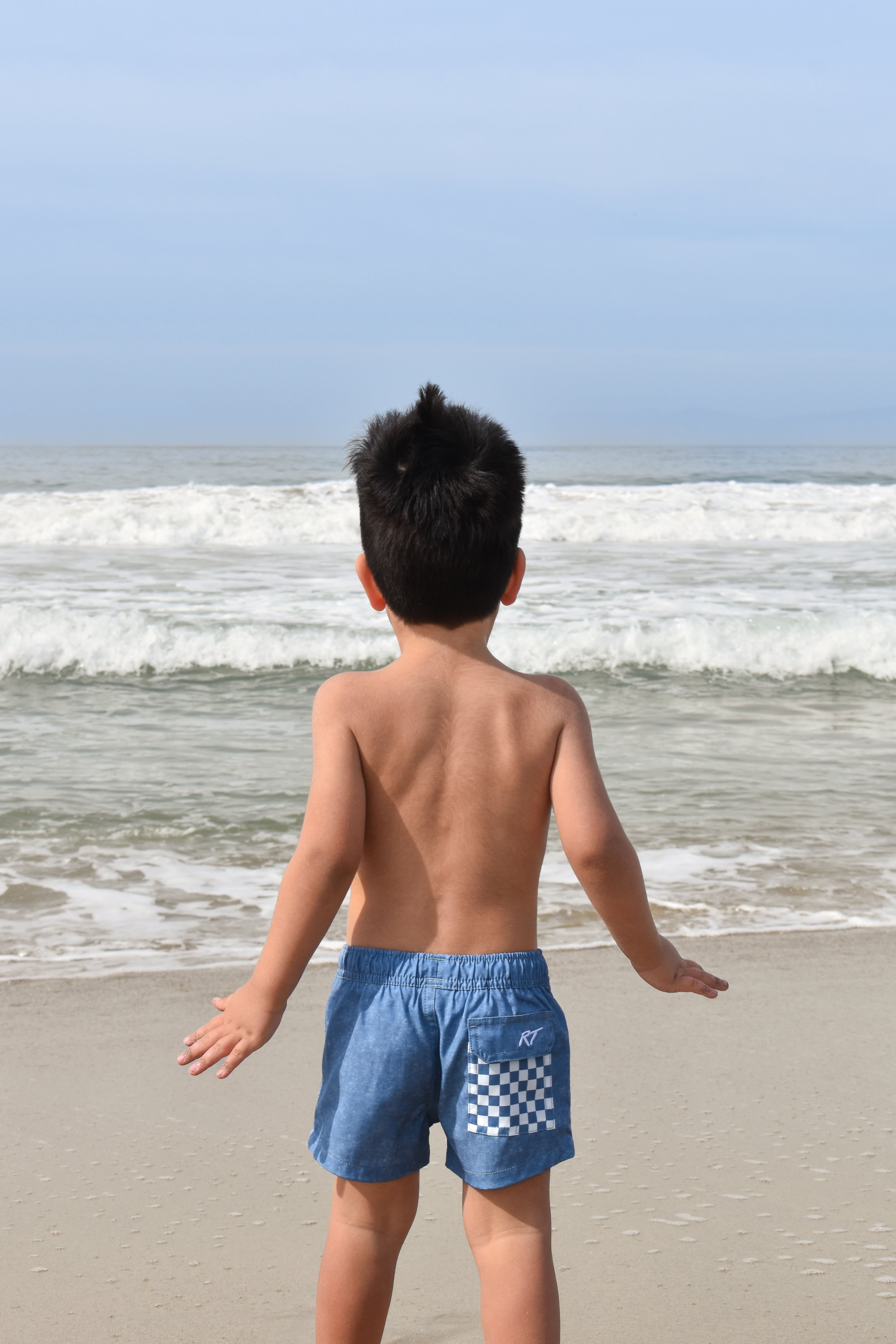 Child wearing blue checkered acid wash shorts standing on a beach with ocean waves in the background by rad toddler