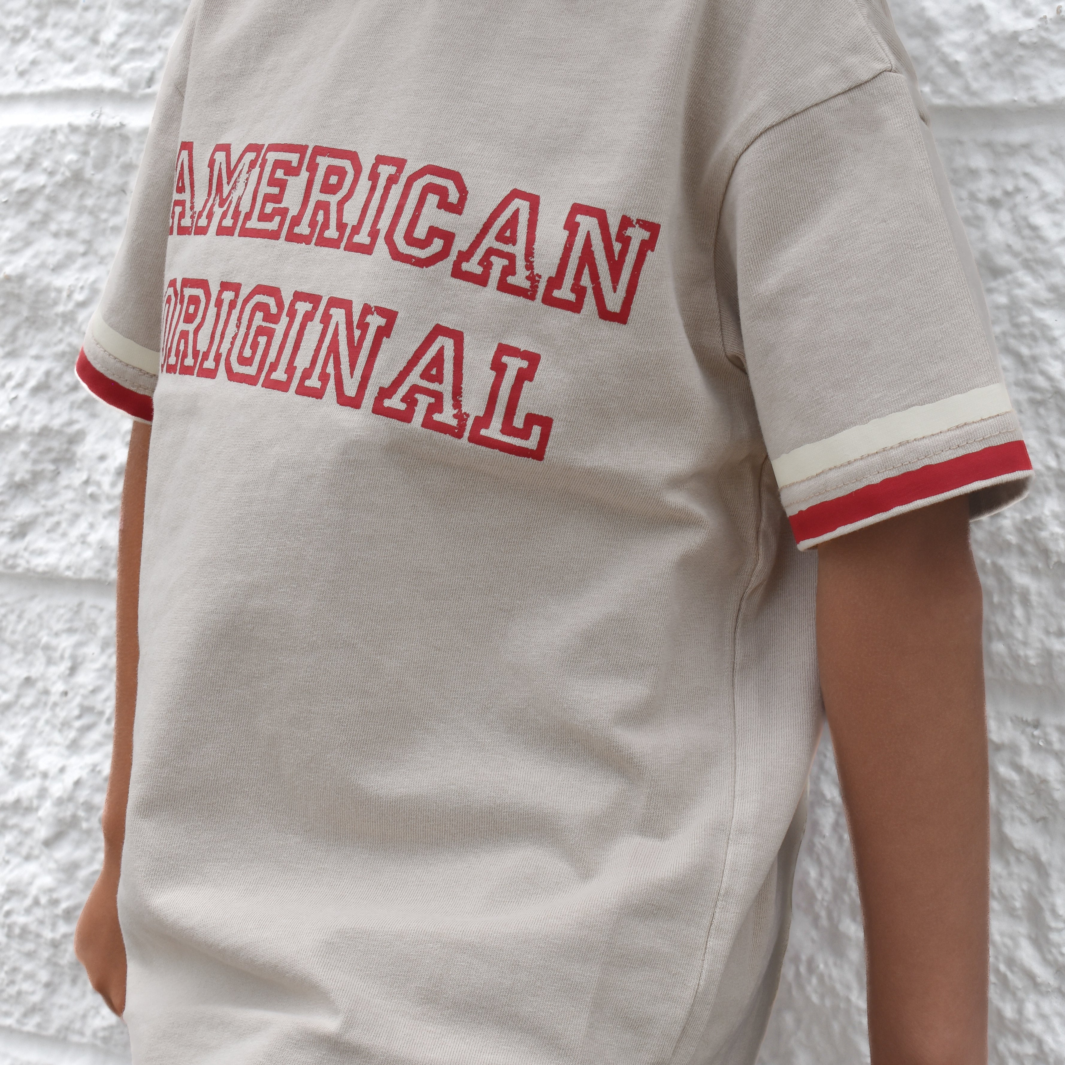 Child wearing a beige t-shirt with 'American Original' text against a textured white wall.