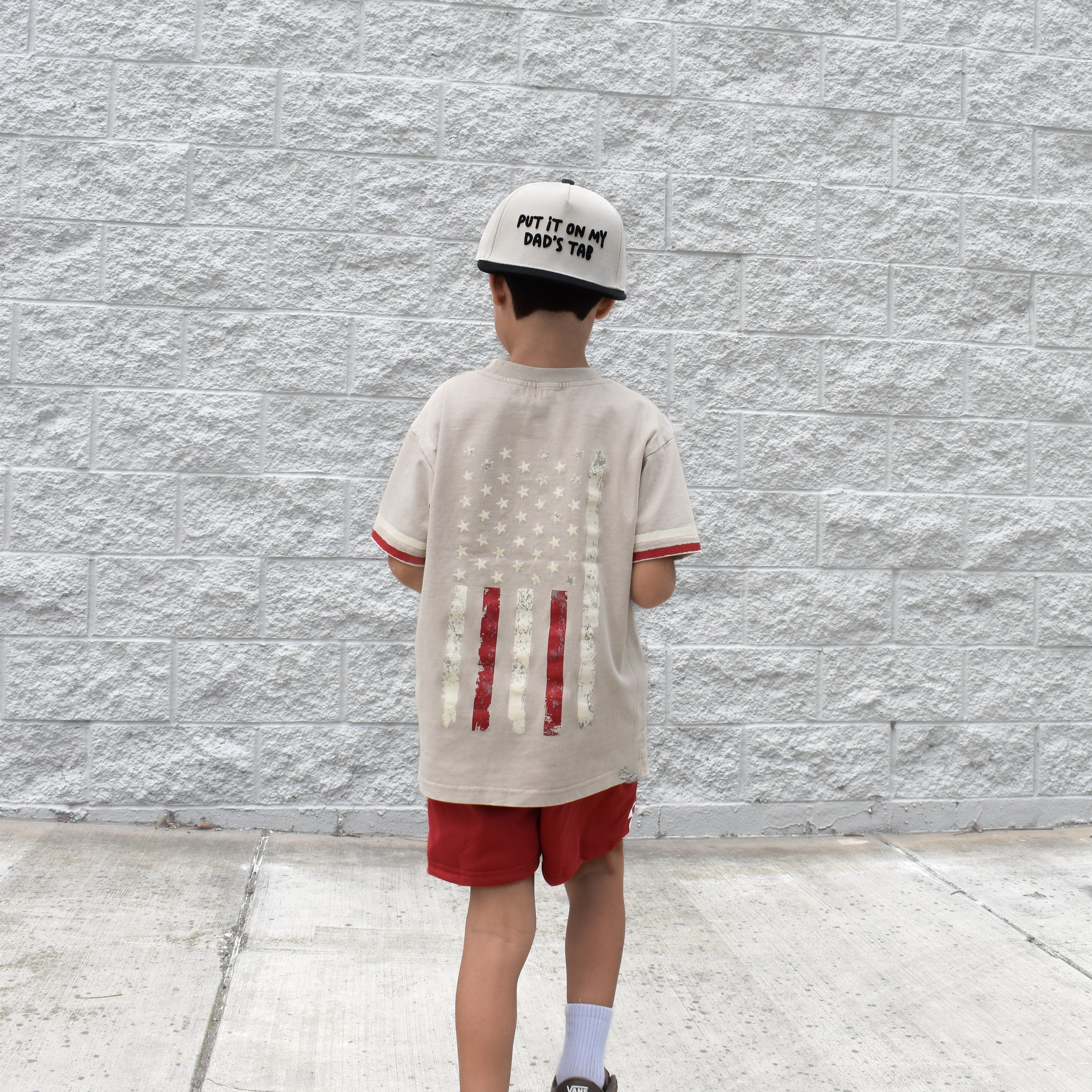 Child wearing a beige t-shirt with red designs and red shorts against a gray textured wall.