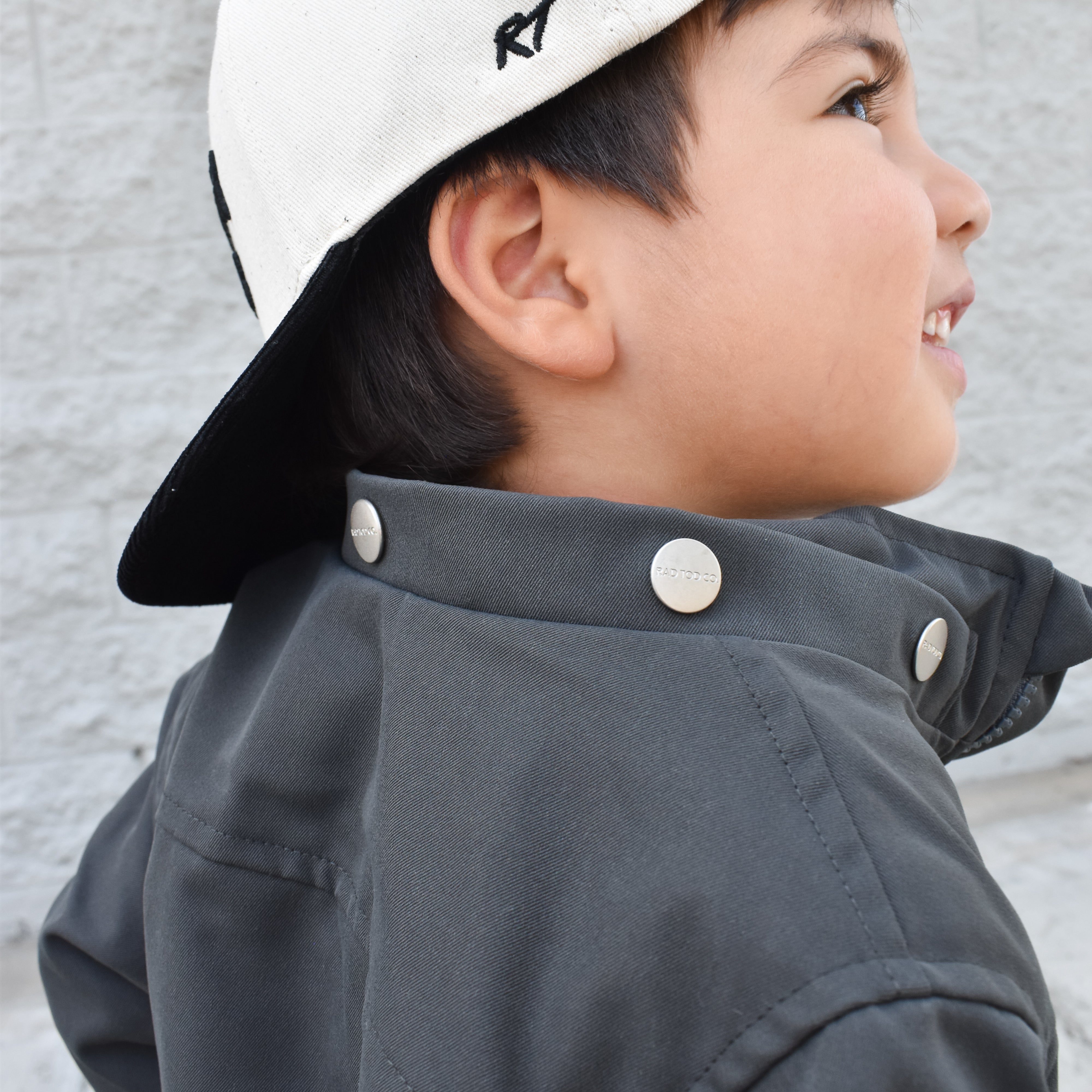 Child wearing a white cap and dark jacket against a light gray background