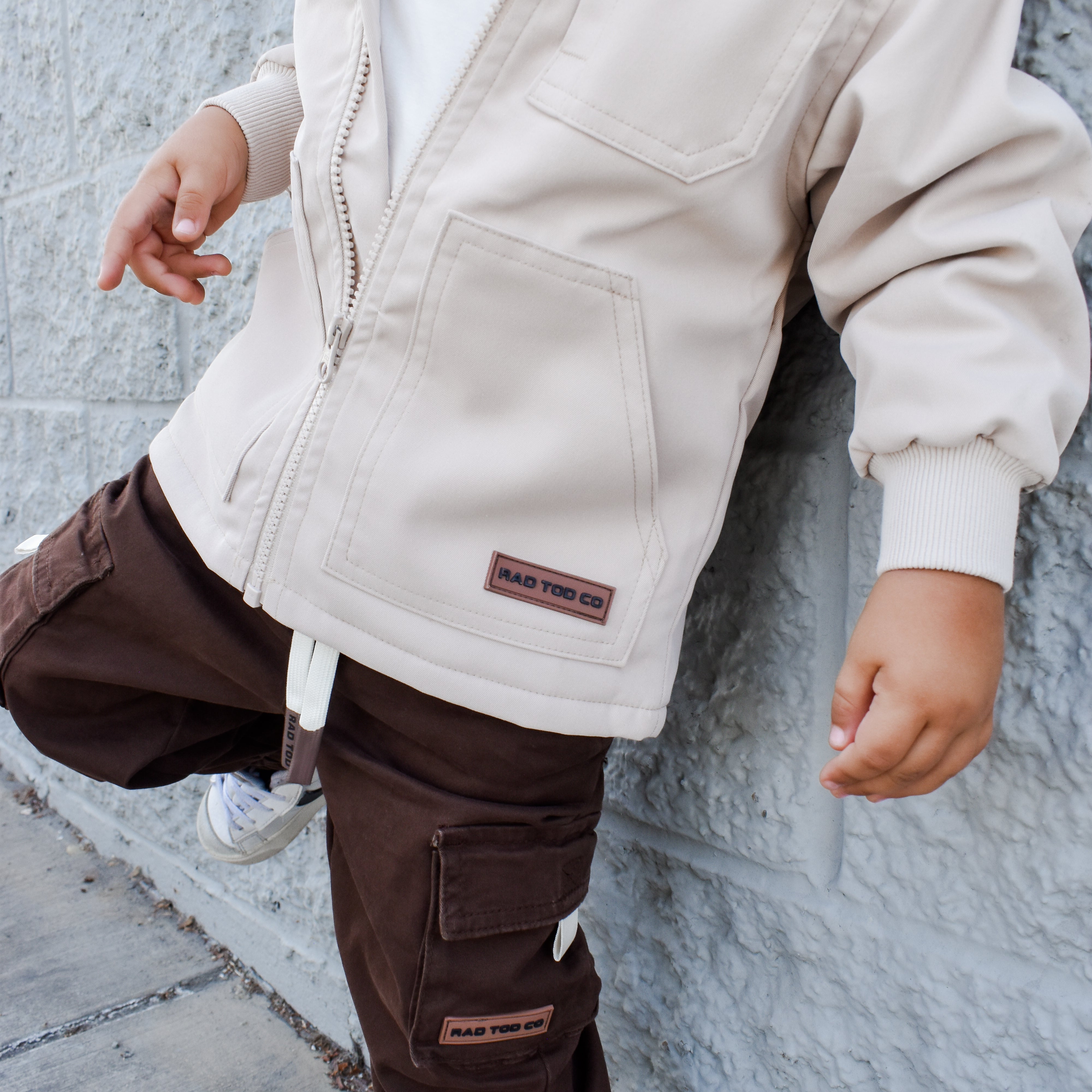 Child wearing a beige jacket and brown pants against a gray wall.