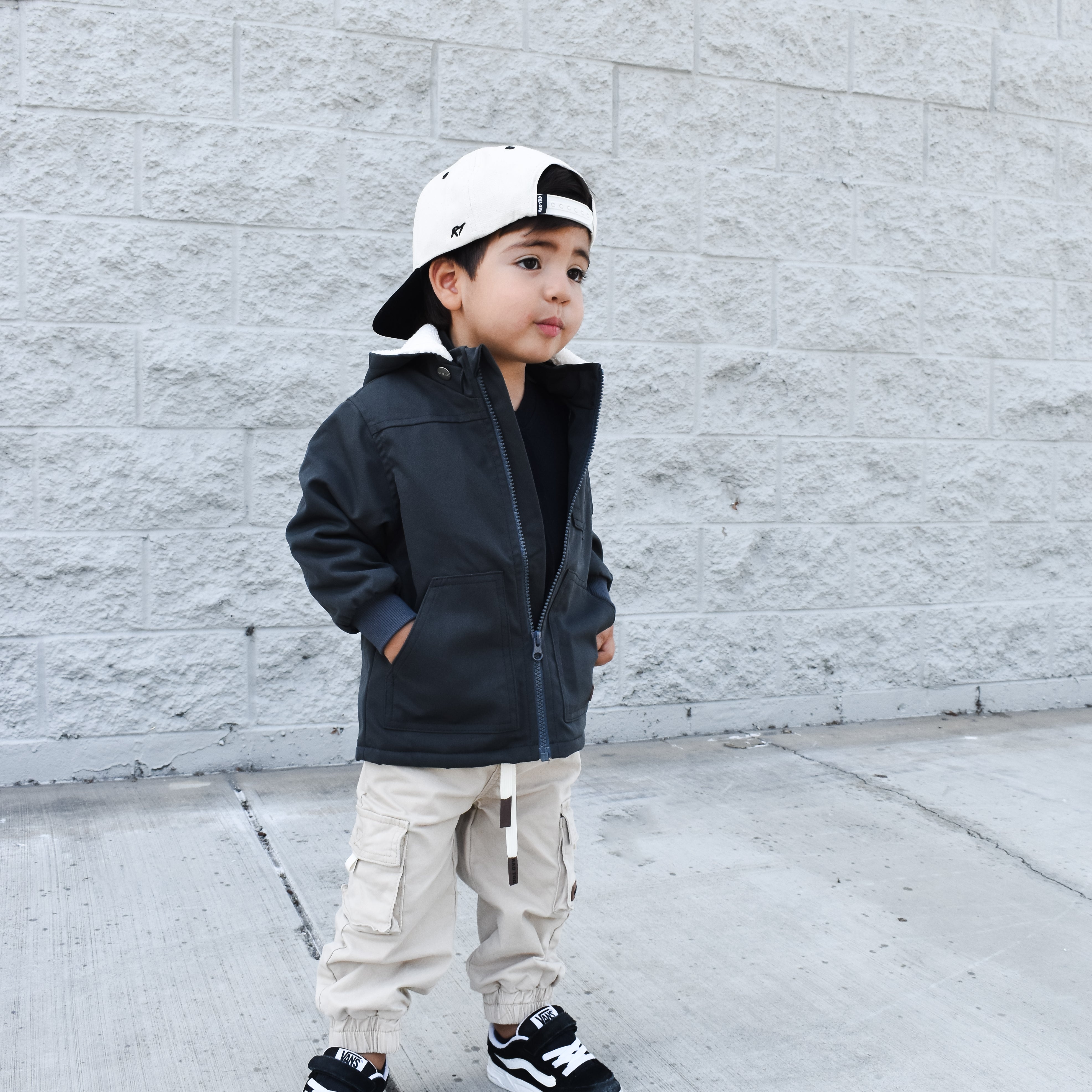 Child wearing a black jacket, beige pants, and a white cap against a gray brick wall.