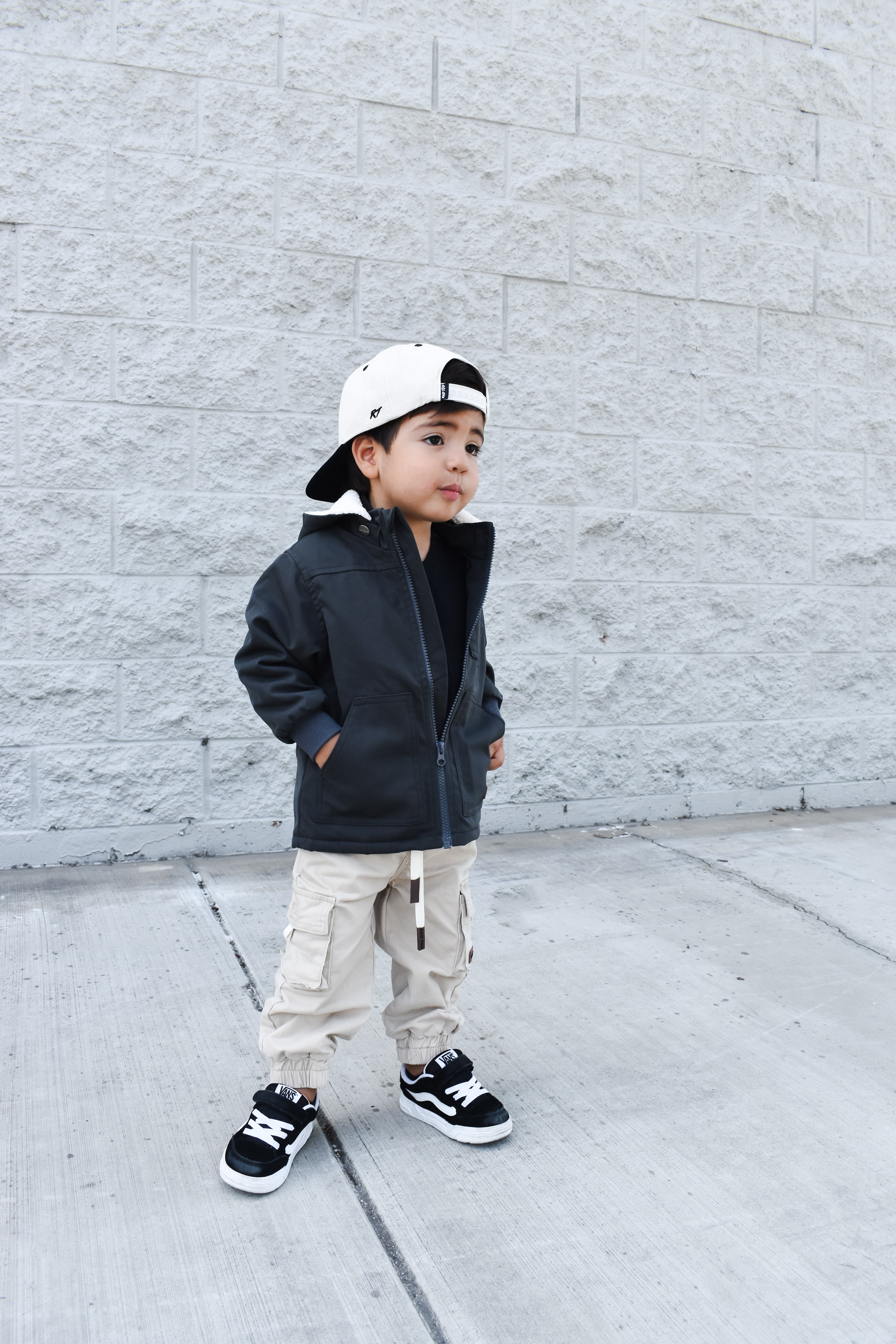 Child wearing a black jacket, beige pants, and a white cap against a gray brick wall.