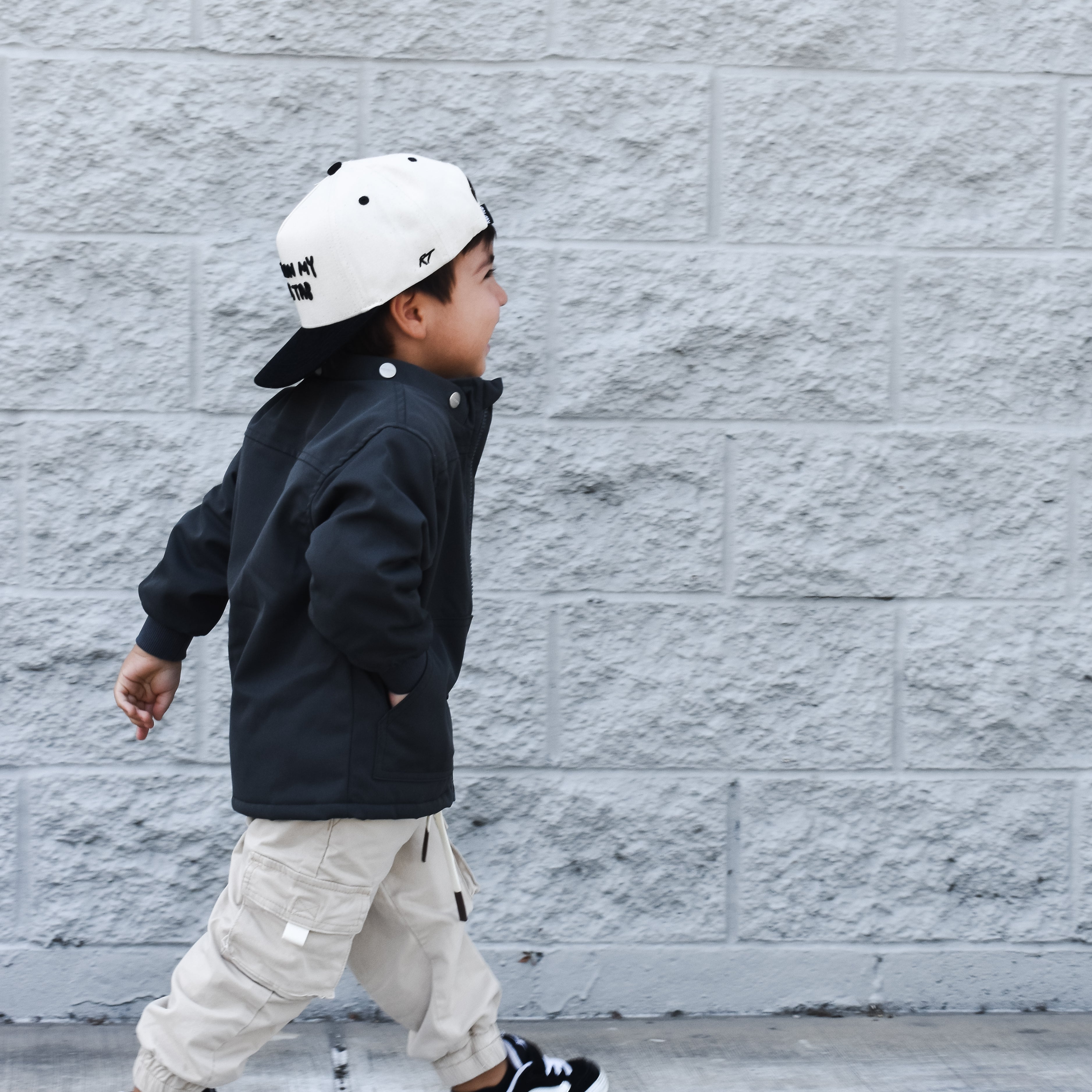 Child wearing a black jacket, beige pants, and a white cap walking against a gray brick wall.