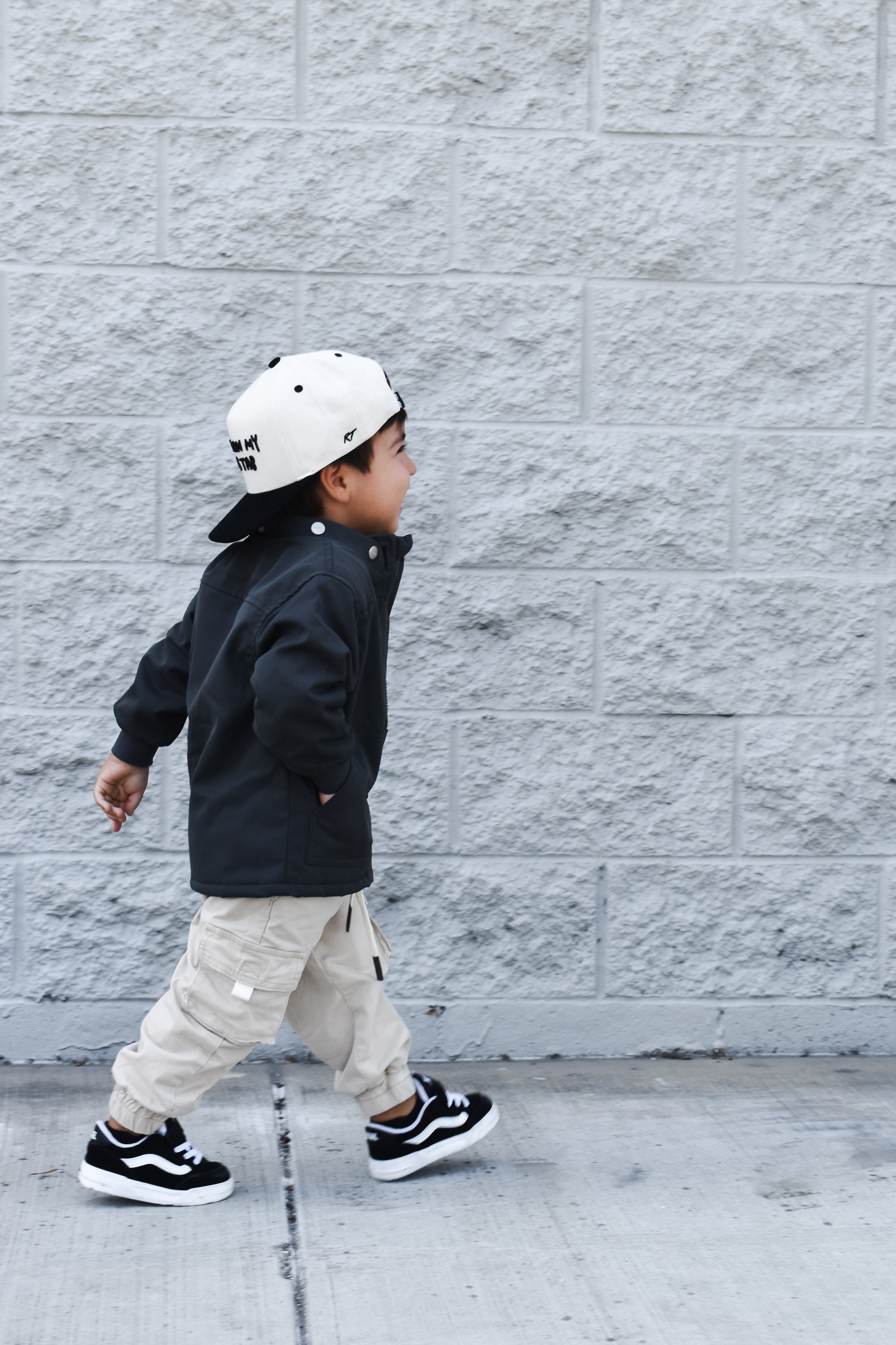 Child wearing a black jacket, beige pants, and a white cap walking against a gray brick wall.