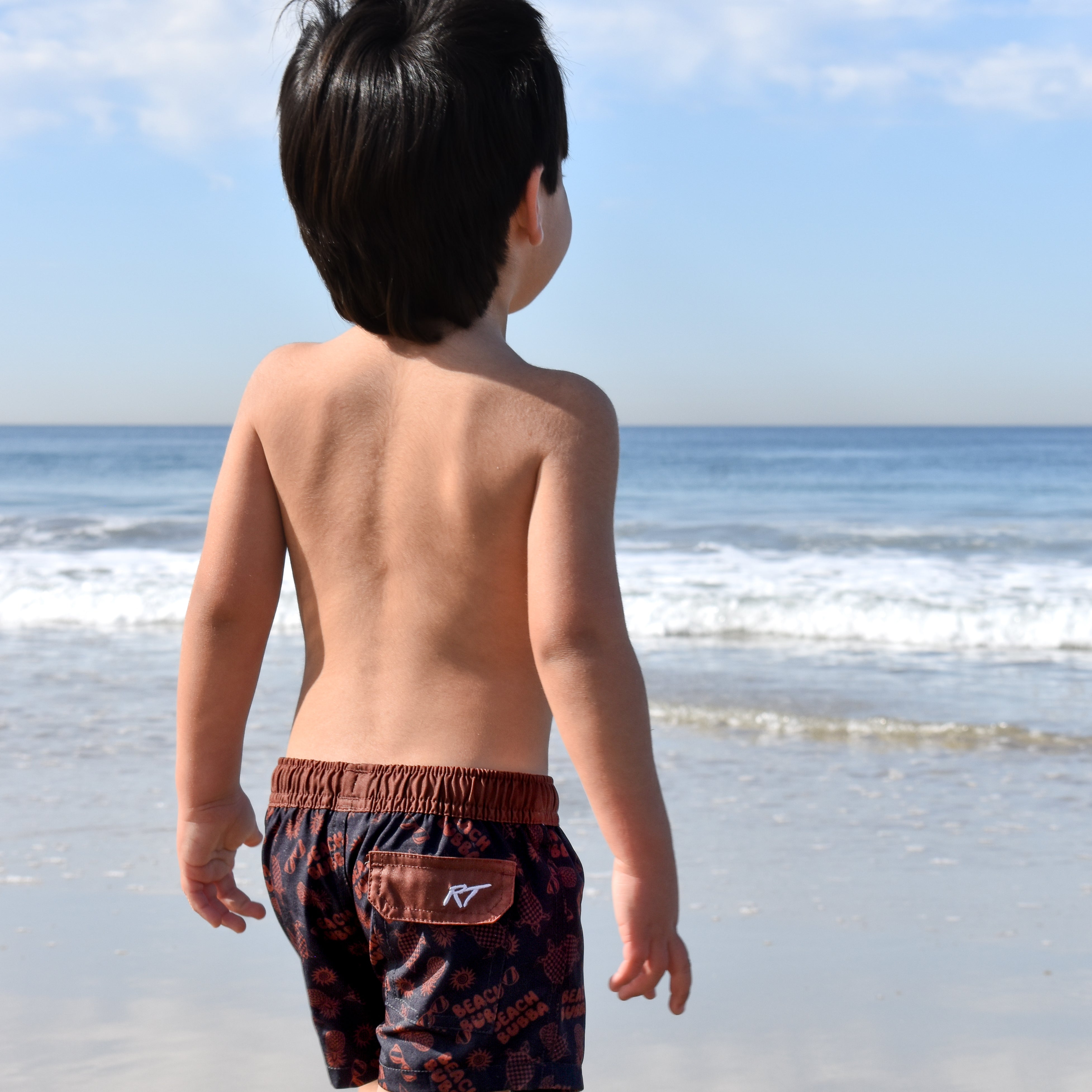 Child wearing swim shorts standing on a beach with ocean and sky in the background