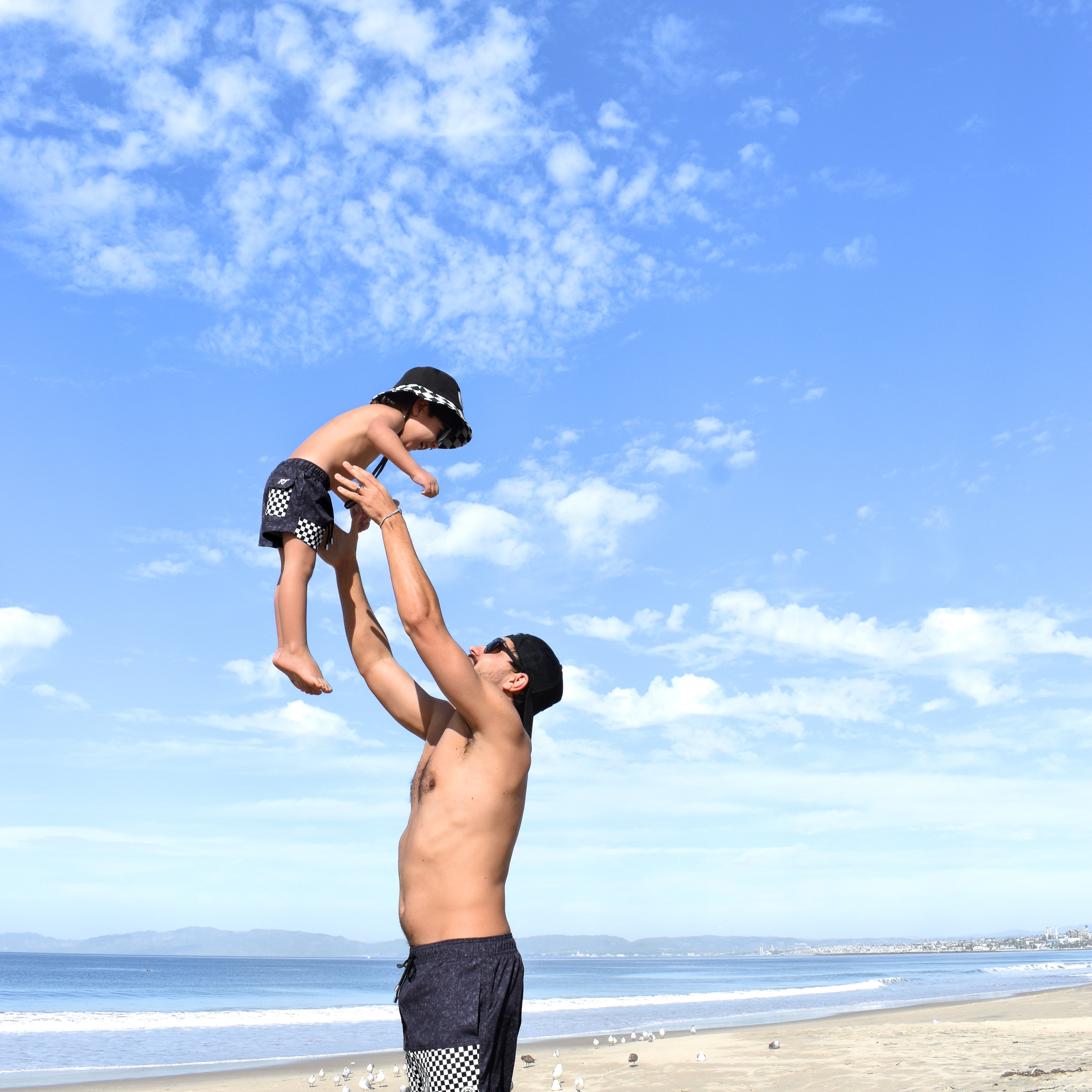 Man lifting a child in the air on a beach with a clear blue sky while sporting some awesome rad toddler gear