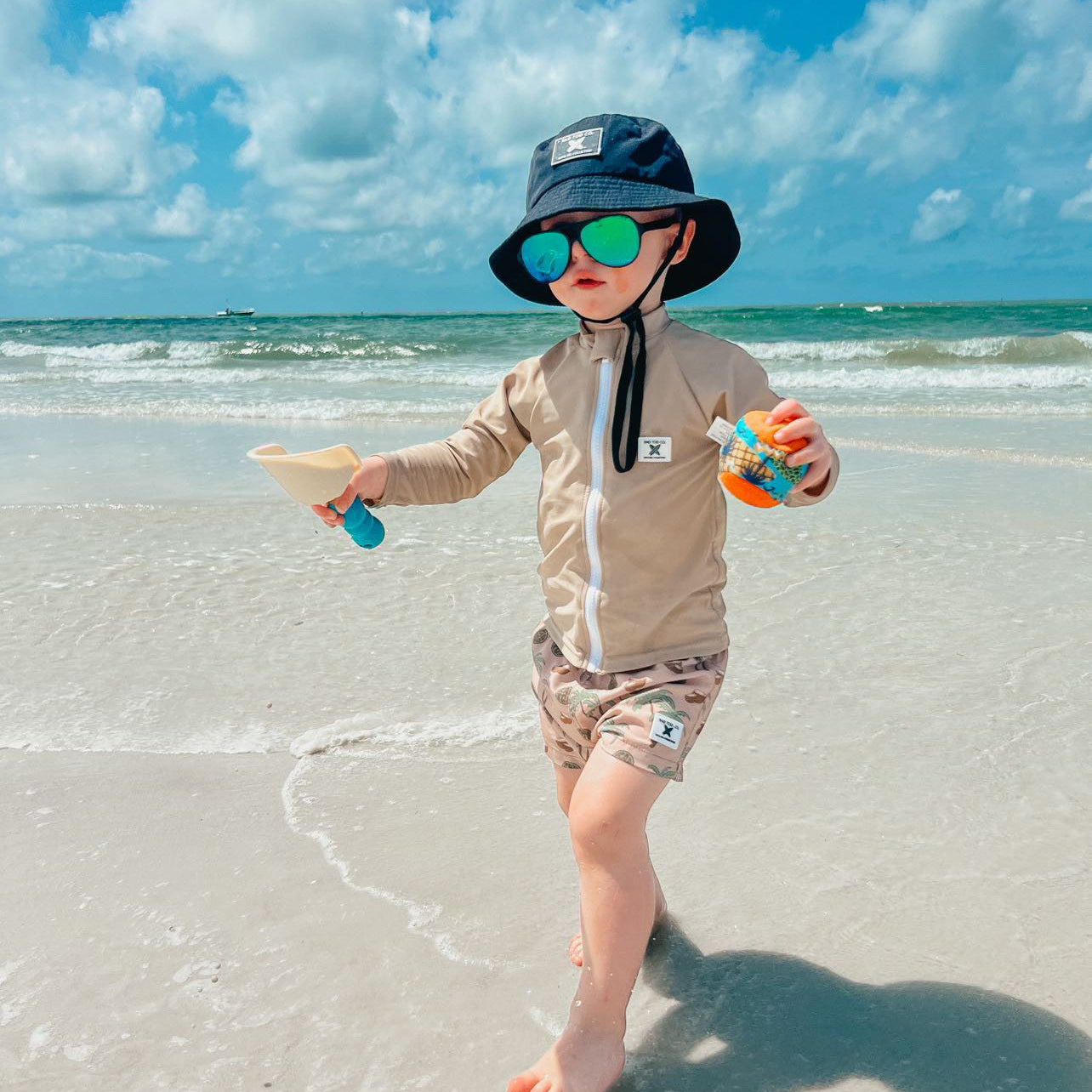 beach day wearing Rad Toddlers 'Sand' Rashguard Shirt with front zipper and UPF 50+ protection, perfect for pairing with board shorts for water activities.