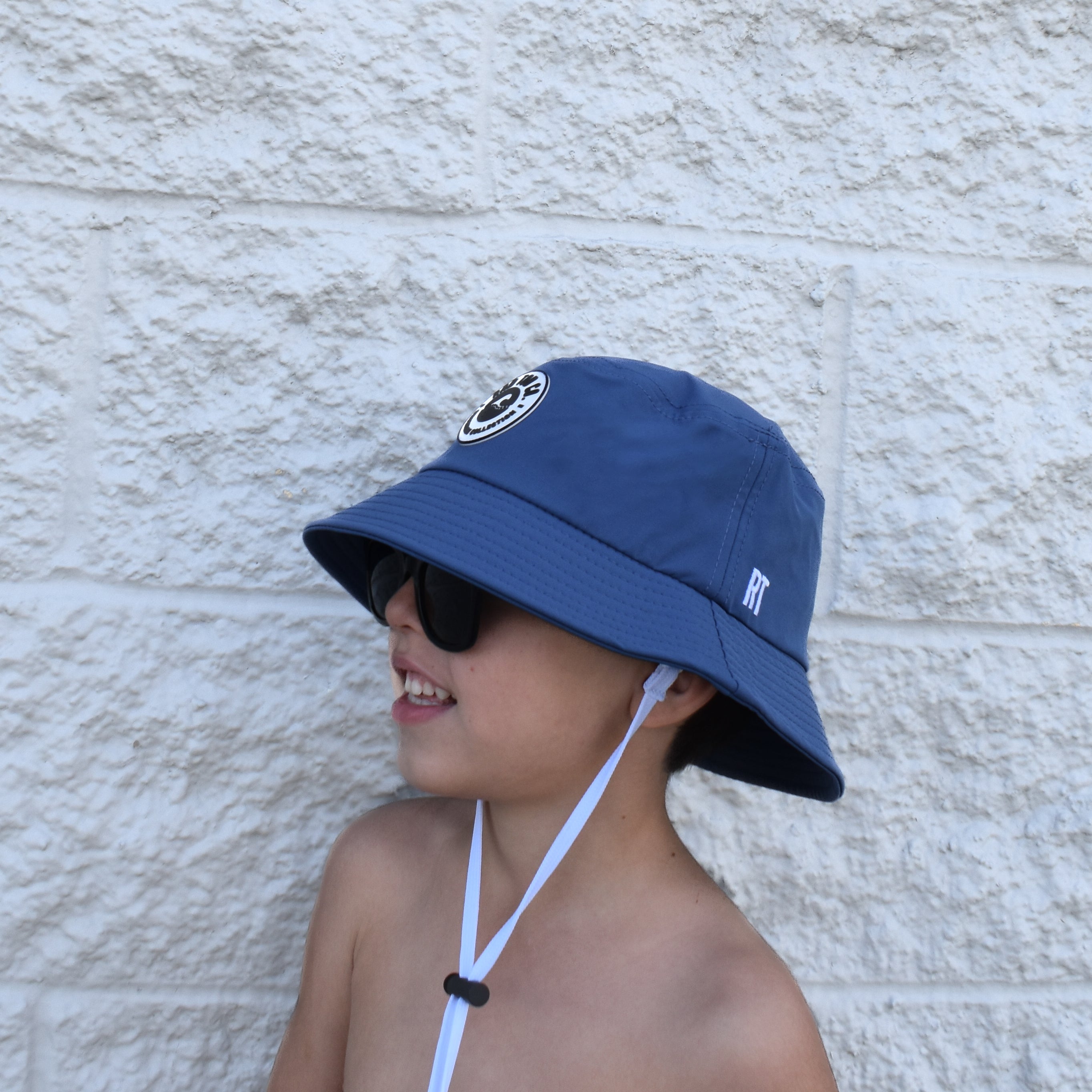 Child wearing a blue bucket hat and sunglasses against a textured wall.