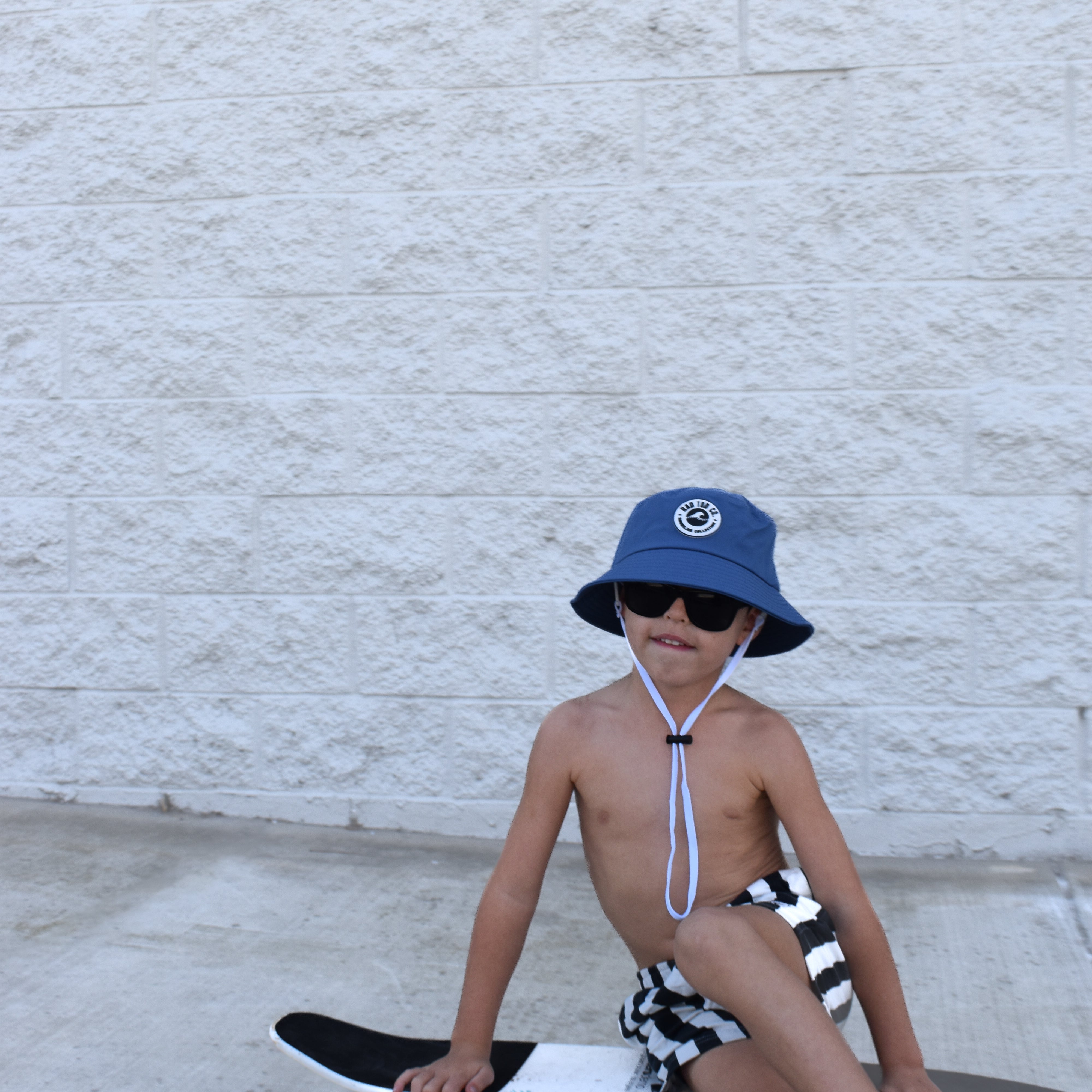 Child sitting on a skateboard wearing a blue bucket hat and sunglasses against a light gray wall.