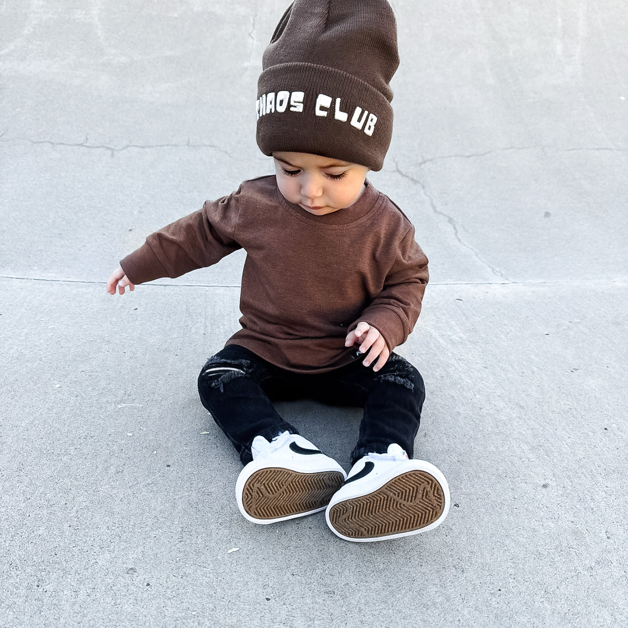 Child wearing a brown outfit with a beanie sitting on a concrete surface