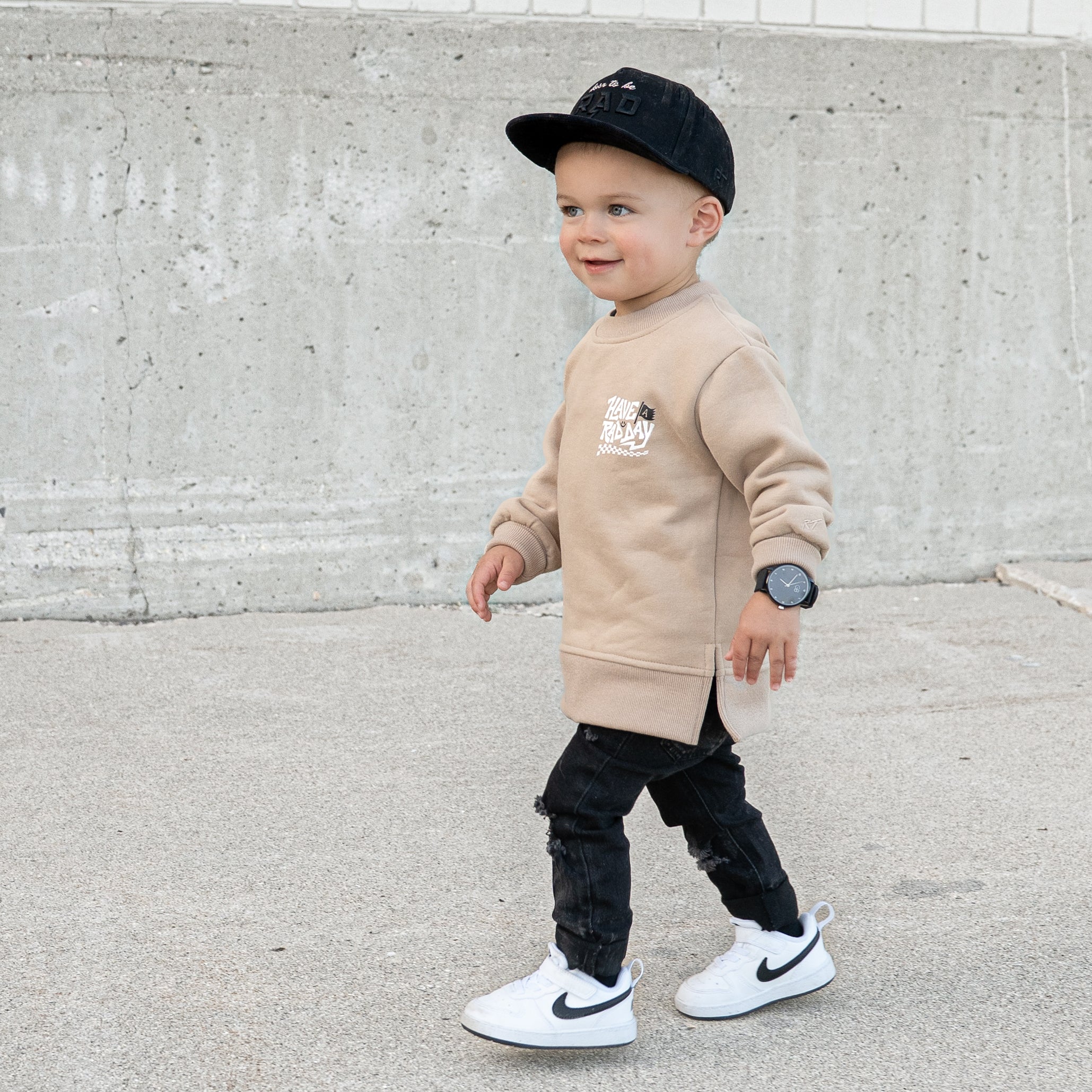 Child wearing beige ‘Have a Rad Day’ crewneck with white lettering, flag, smiley face, and checkers
