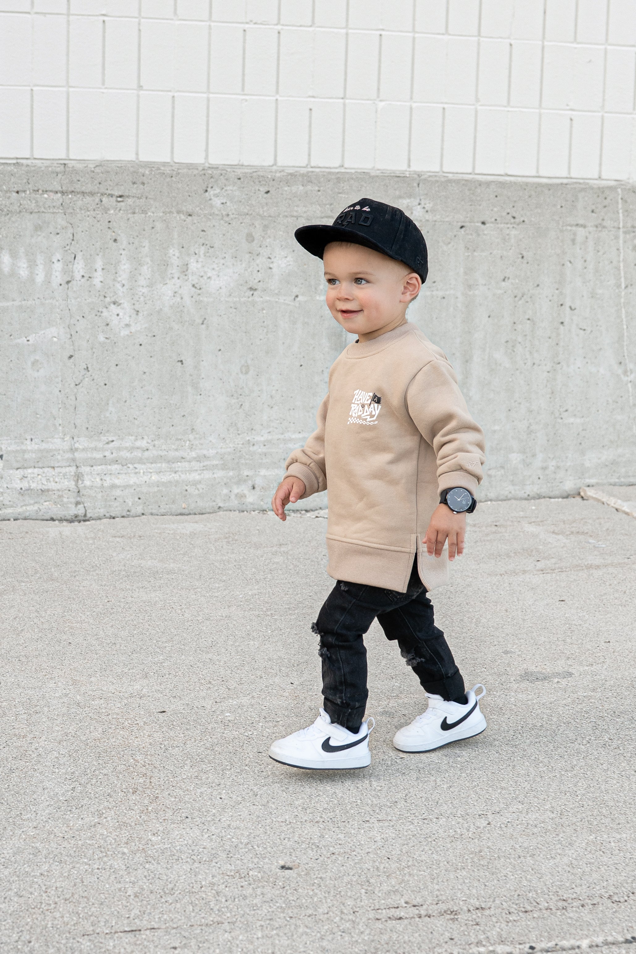 Child wearing beige ‘Have a Rad Day’ crewneck with white lettering, flag, smiley face, and checkers