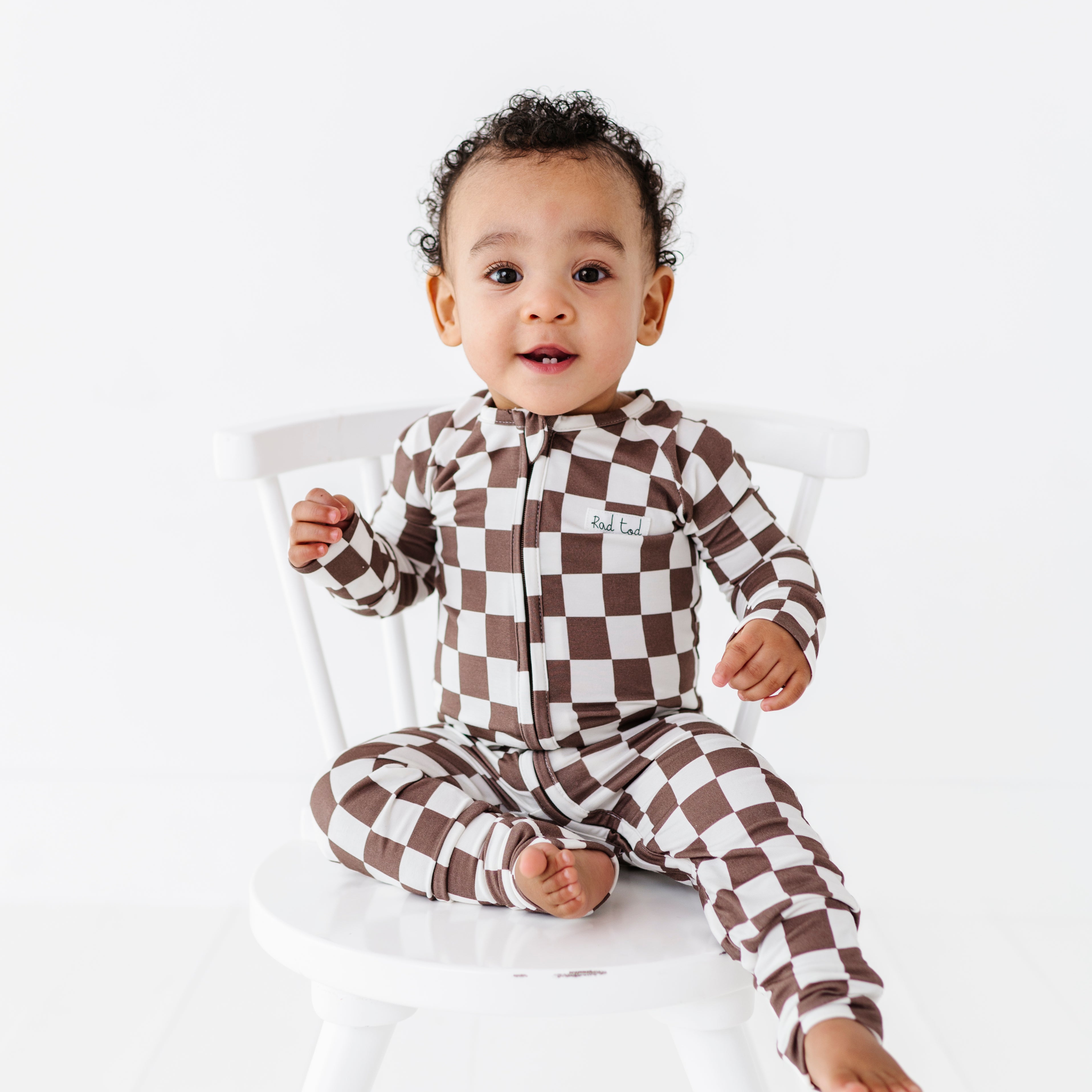 boy sitting on chair wearing Bronze and white checkered bamboo pajamas for toddlers, featuring a two-way zipper and foldover footies by Rad Toddler.