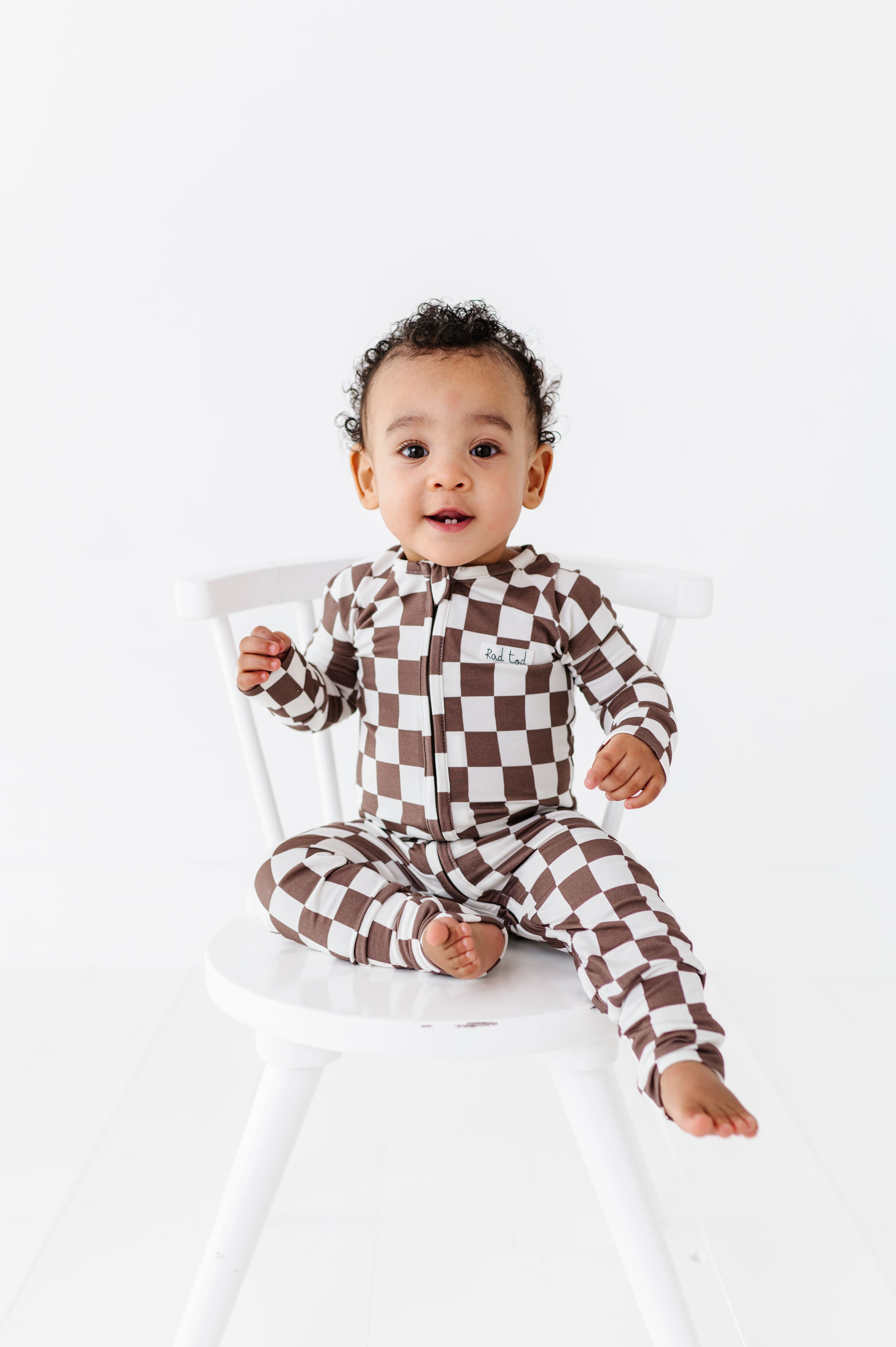 boy sitting on chair wearing Bronze and white checkered bamboo pajamas for toddlers, featuring a two-way zipper and foldover footies by Rad Toddler.