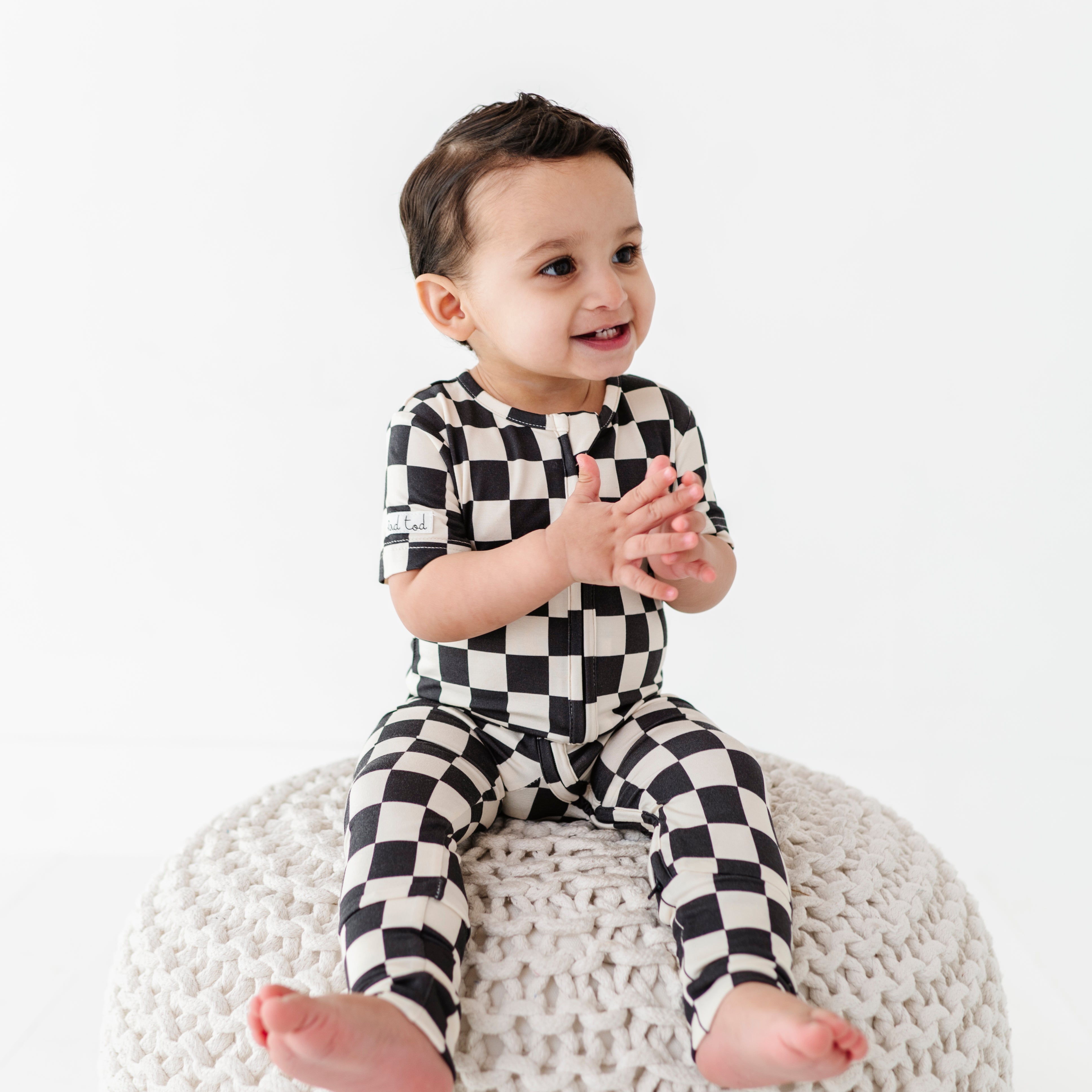 boy sitting while wearing Black and white checkered bamboo short sleeve onesie for toddlers, featuring a two-way zipper and optional foldover footies by Rad Toddler.