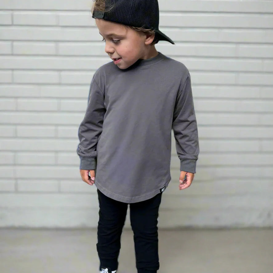 Child wearing a gray long-sleeve shirt, black pants, and black sneakers with a cap against a light gray background.
