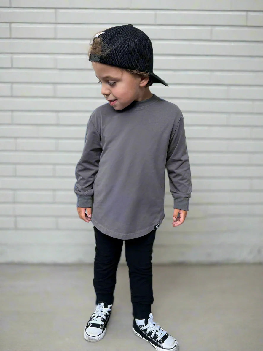 Child wearing a gray long-sleeve shirt, black pants, and black sneakers with a cap against a light gray background.