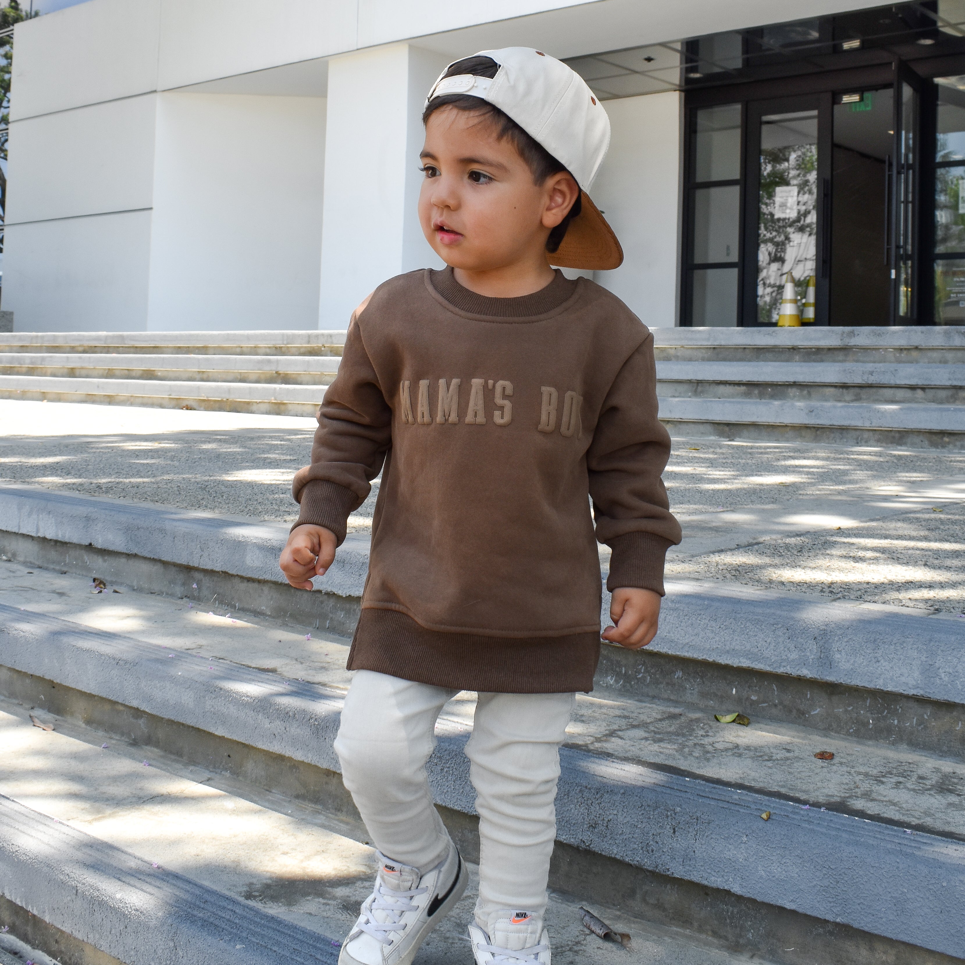 Child wearing brown “Mama’s Boy” crewneck with lighter brown lettering, relaxed fit, paired with casual bottoms.
