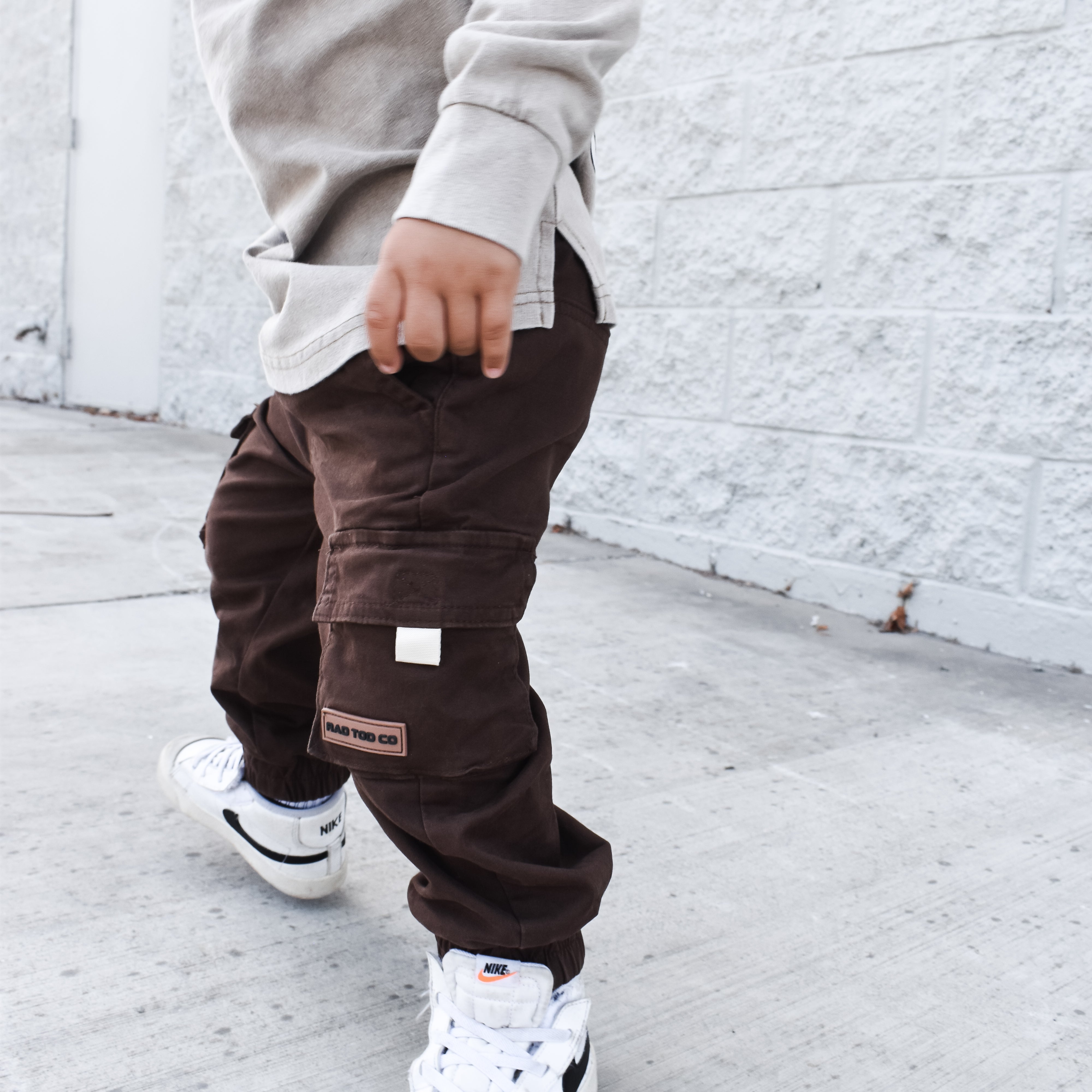 Child wearing a gray sweatshirt and brown cargo pants with white sneakers against a white brick wall by RAD TODDLER
